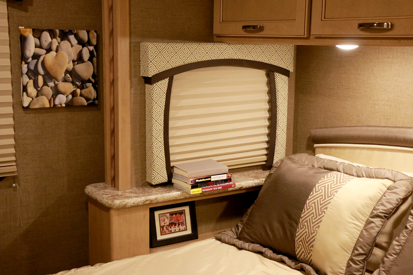 Cozy RV rental bedroom featuring a bed with pillows, a window with a patterned valance, bedside shelf, books, and art.. Thor Motor Coach Chateau 2018