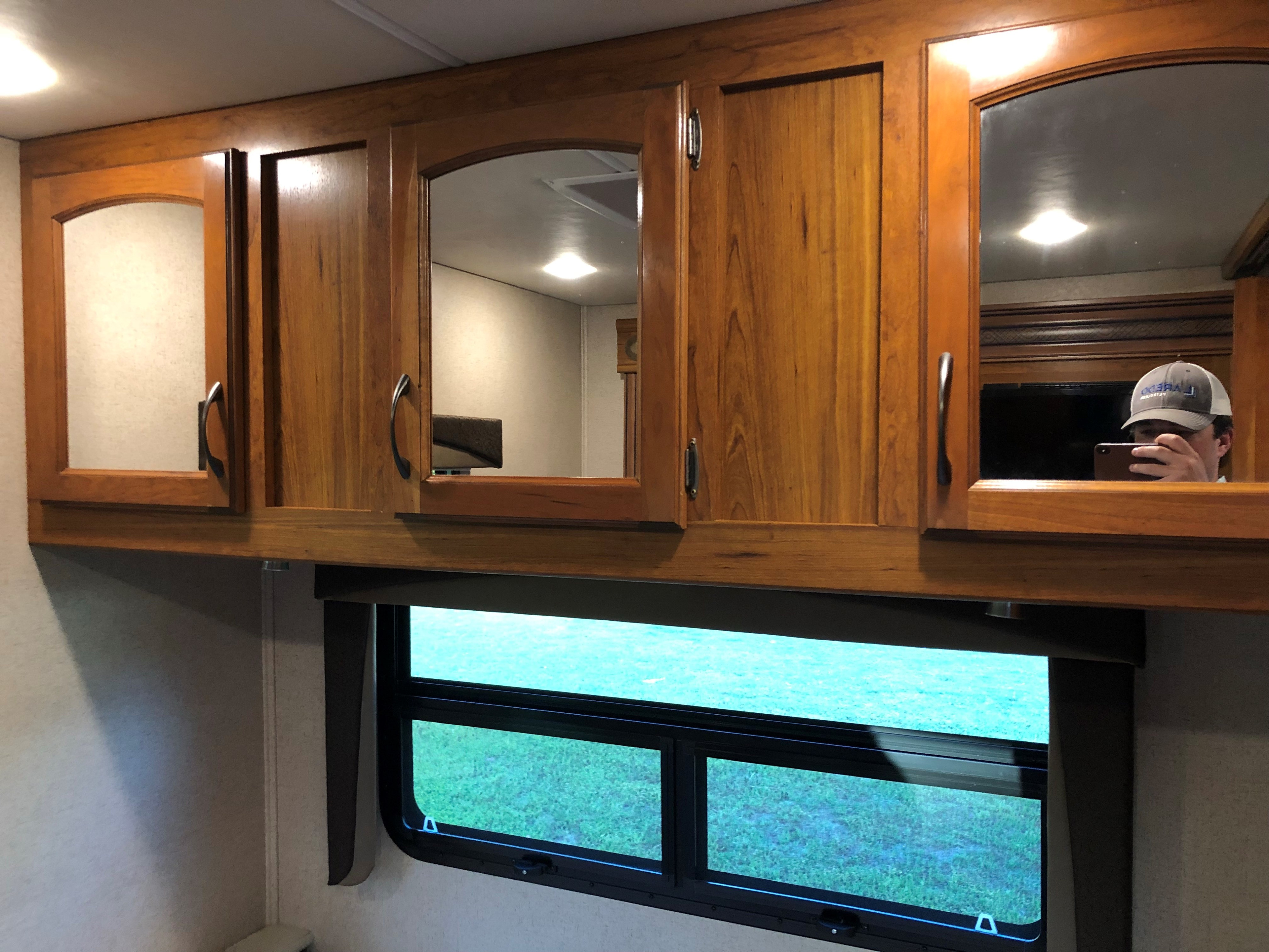RV rental: wooden overhead cabinets, some with mirrors showing a reflection of a person. Below, a window looks out onto green grass.. Jayco Greyhawk 2016