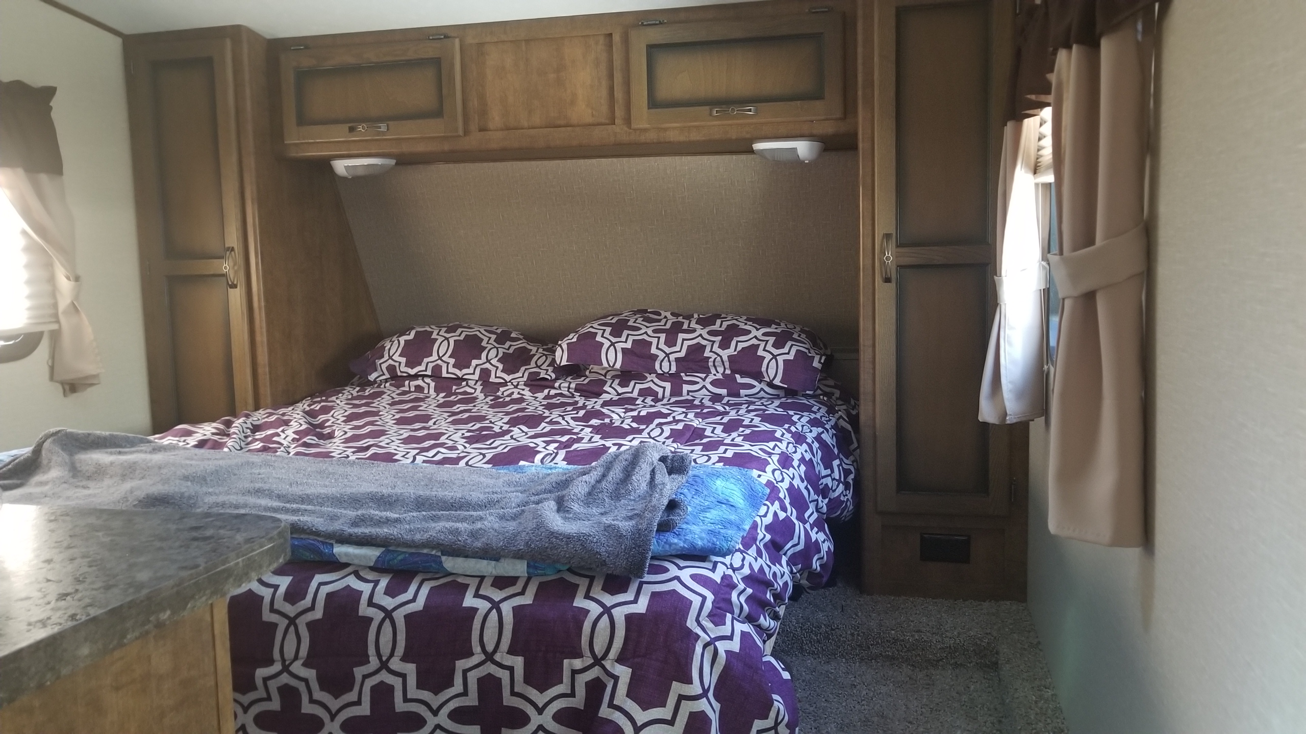 Comfortable RV rental bedroom showing a queen bed with purple and white bedding, overhead cabinets, and side closets.. Crossroads RV Rezerve 2016