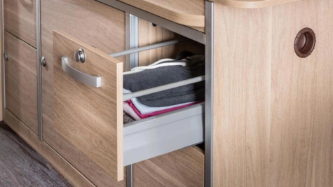 A wooden cabinet in an RV rental, with a partially open drawer revealing folded clothes & a grey storage bin.. Hymer Aktiv 2019