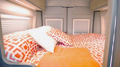 RV rental bedroom: a cozy space with vibrant orange patterned bedding, white pillows, and windows with blinds, providing comfort on the go.. Hymer Aktiv 2019