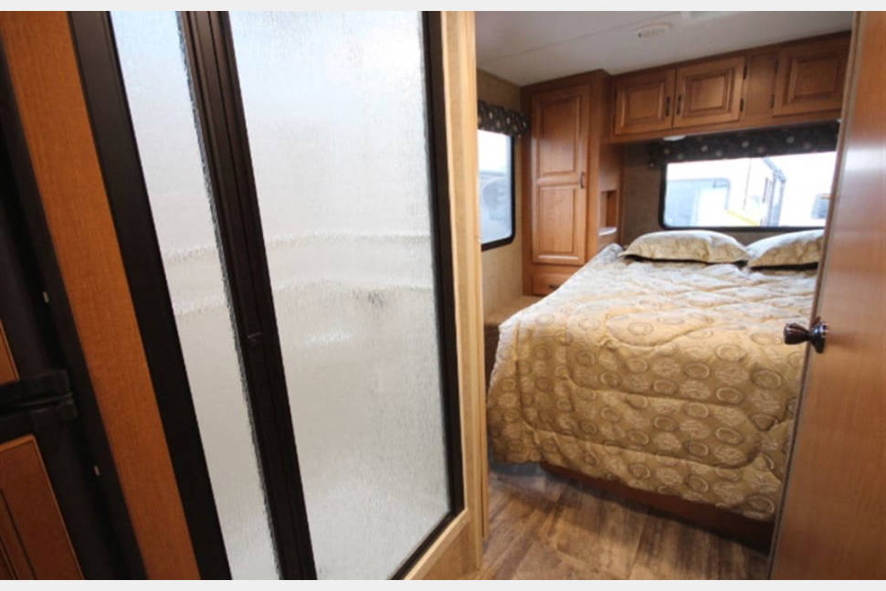 Inside an RV rental: a compact bedroom with a queen bed and overhead cabinets, adjacent to a modern frosted glass shower.. Forest River Sunseeker 2017