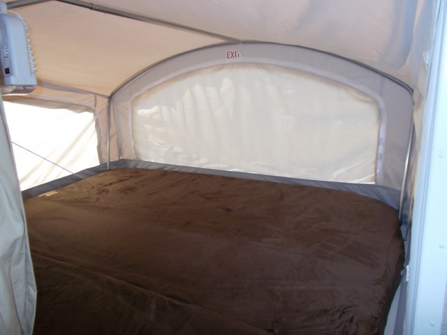 Inside a cozy RV rental pop-up camper: a comfy, large bed with a brown blanket and a small fan nearby.. Keystone Bullet 2018