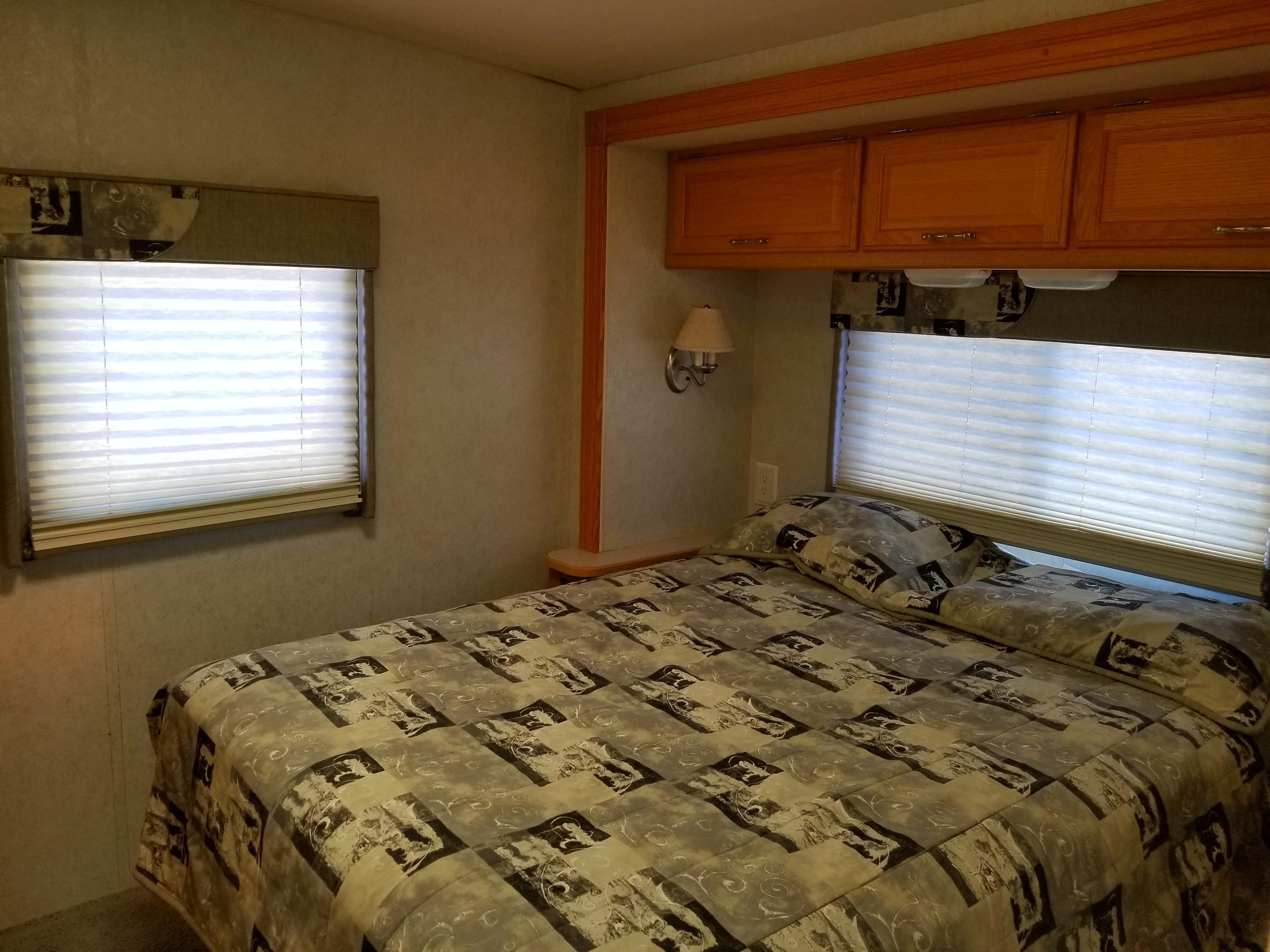 Cozy RV rental bedroom featuring a patterned bed, window blinds, wall sconce, and overhead cabinets.. Winnebago Outlook 2006