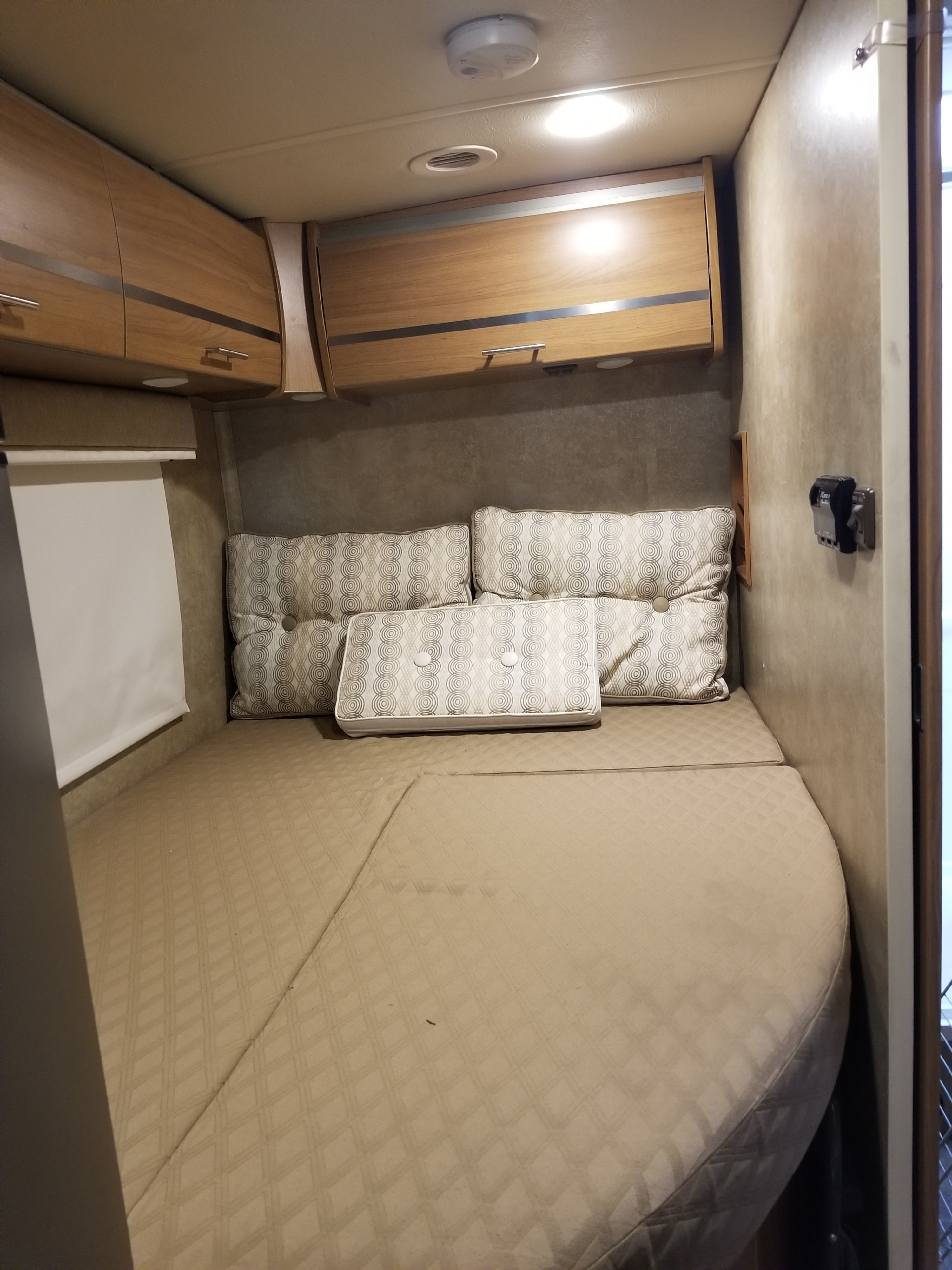 Inside an rv rental, a comfortable bed with quilted mattress and patterned pillows. Overhead wood cabinets and lighting provide storage.. Itasca Reyo 2014