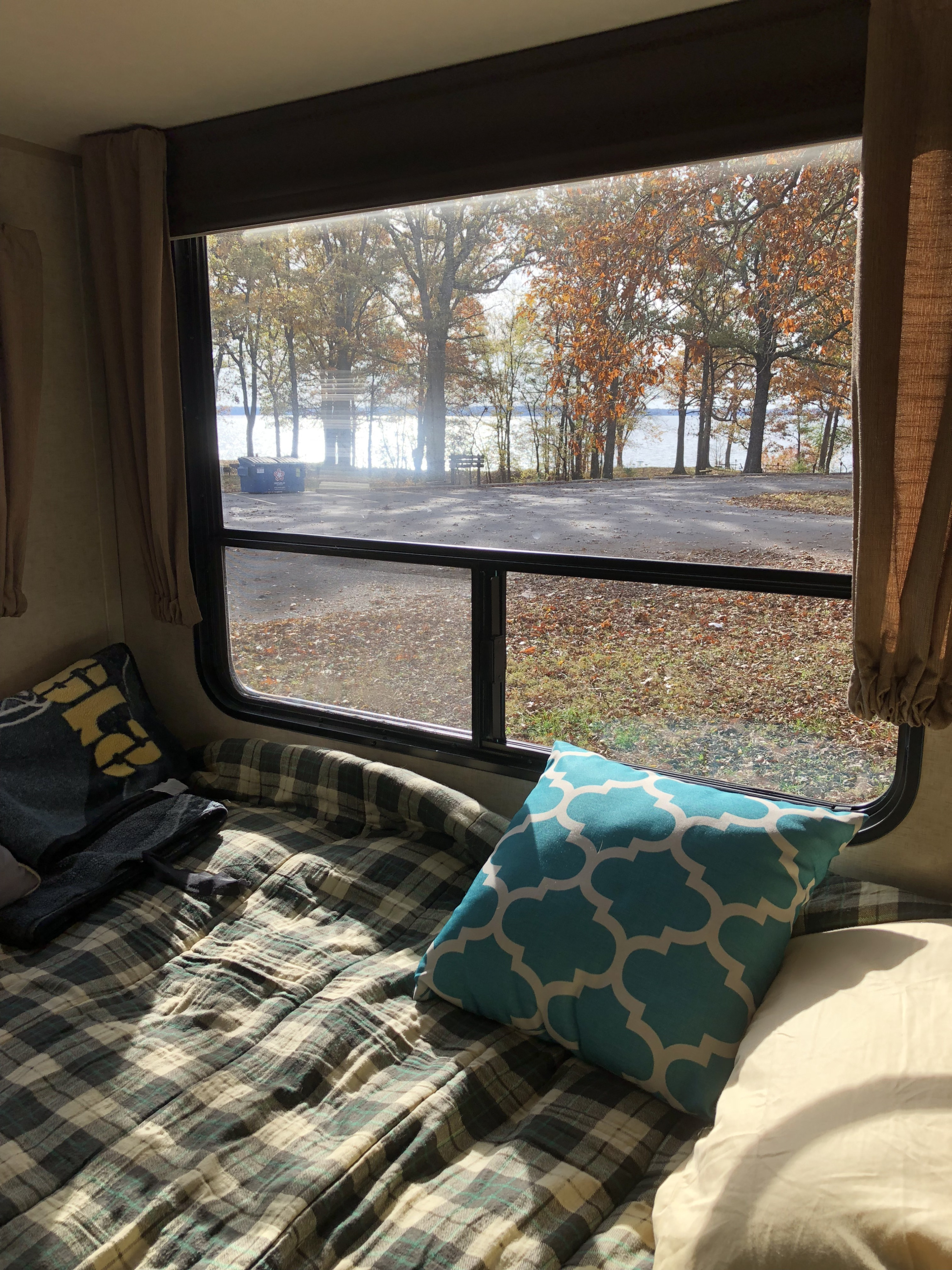 Enjoy the view from this cozy RV rental! A bed with plaid bedding and a blue pillow overlooks an autumn lake scene.. Forest River Cherokee 2018