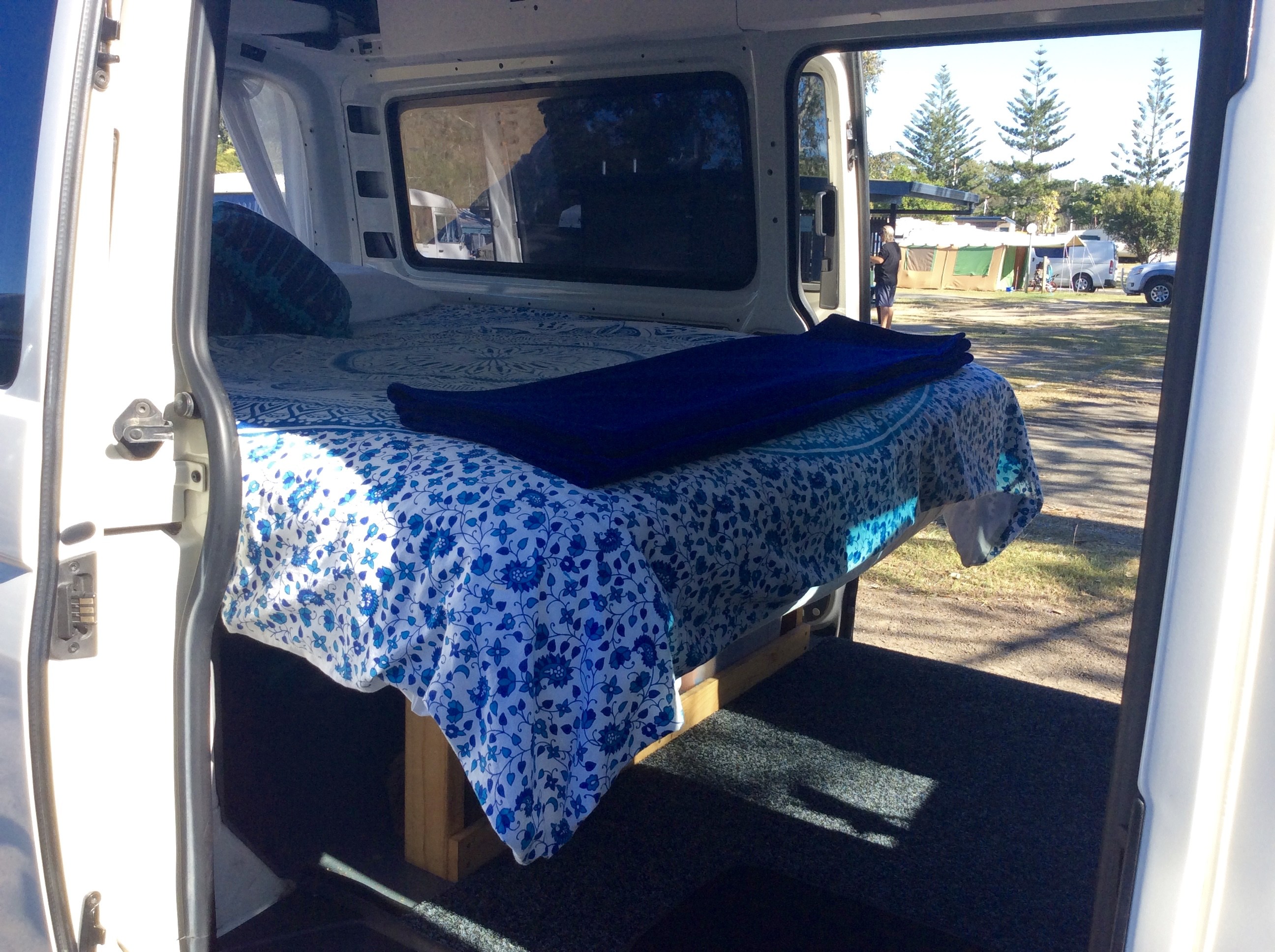 Comfortable RV rental bed inside a camper van, open to a sunny campsite view. Ideal for road trip adventures.. Volkswagen Transporter 2012