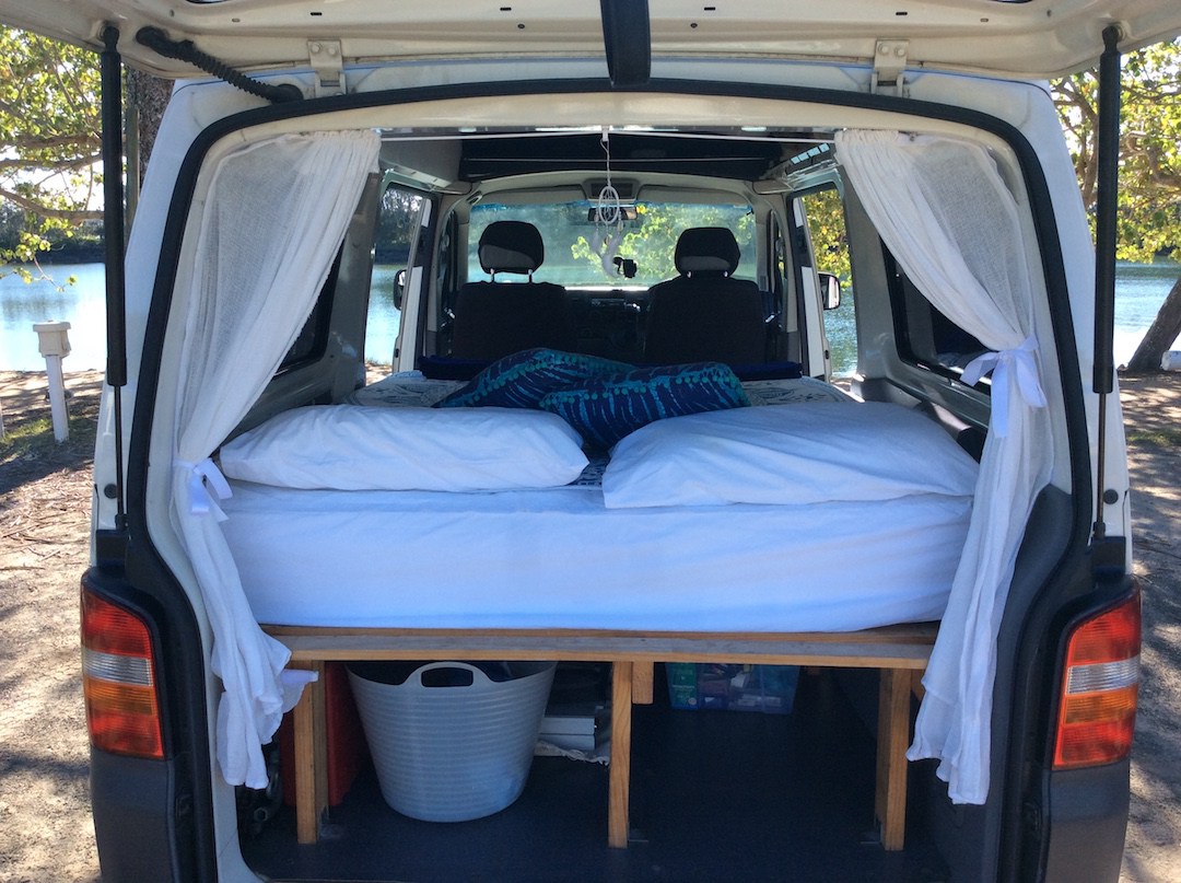 Inside an RV rental: a cozy van bed with white linens, curtains, and a beautiful view of water and trees. Ideal for travel.. Volkswagen Transporter 2012