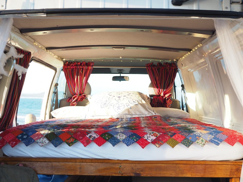 Dreamy camper van bedroom ready for your RV rental adventure. Vibrant quilt, string lights, and ocean view await.. Mitsubishi Express 2006