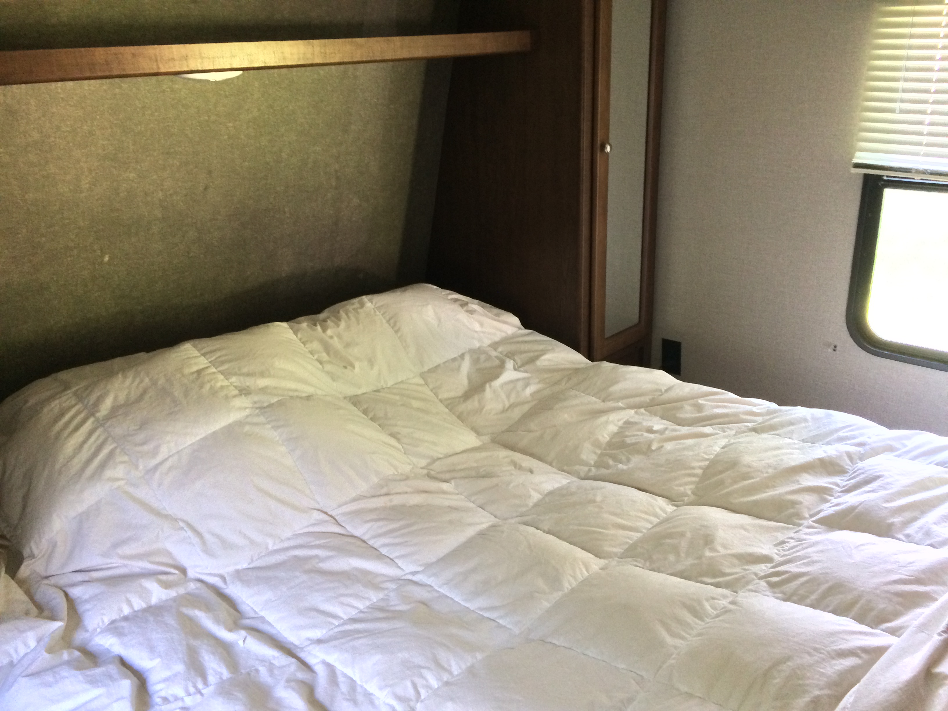 Cozy RV rental bedroom with a white comforter on the bed, overhead shelf, and window with blinds.. Keystone Hideout 2017