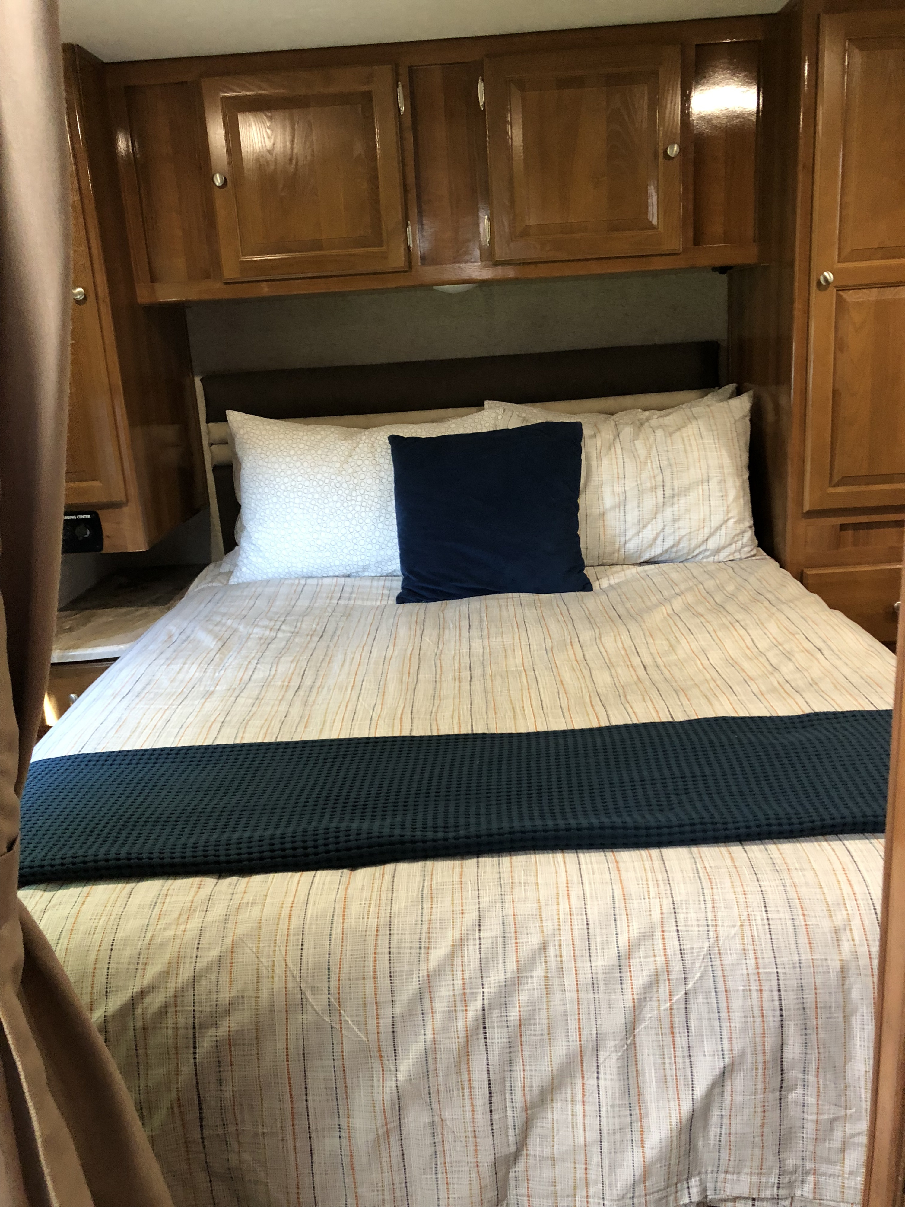 Comfortable RV rental sleeping area: striped bedding, blue pillows, and overhead wooden cabinets.. Forest River Coachmen Leprechaun 2019