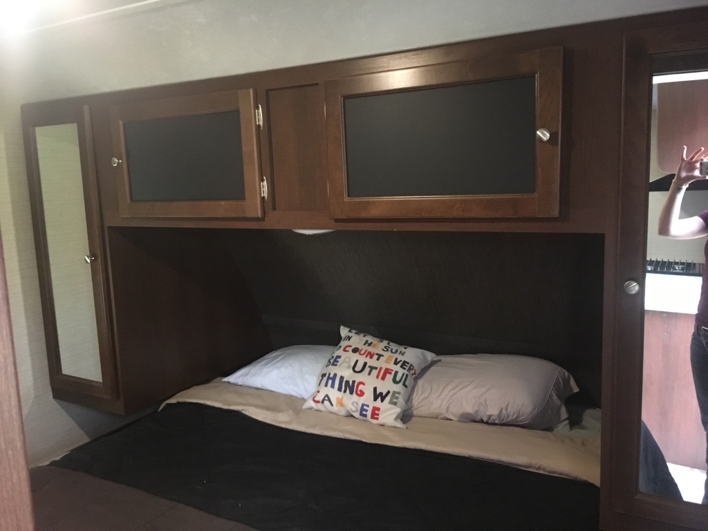 RV rental compact bedroom: a cozy bed with pillows, dark wood overhead cabinets, and a long mirror on the left.. Keystone RV Bullet Ultra Lite 2016
