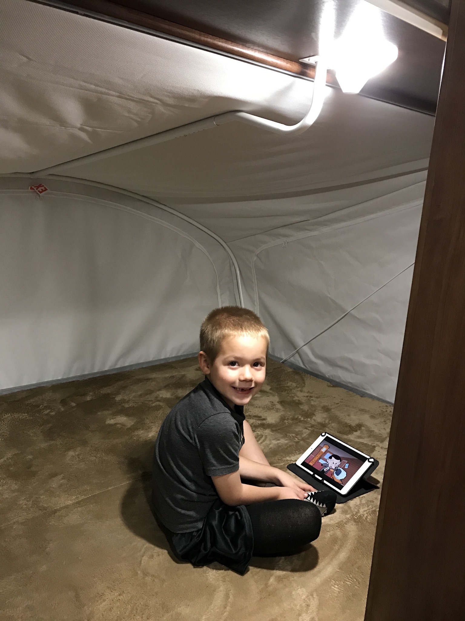 A smiling child relaxes in a cozy RV rental pop-out bunk, watching a tablet on the carpeted floor.. Jayco Jay Feather 2017