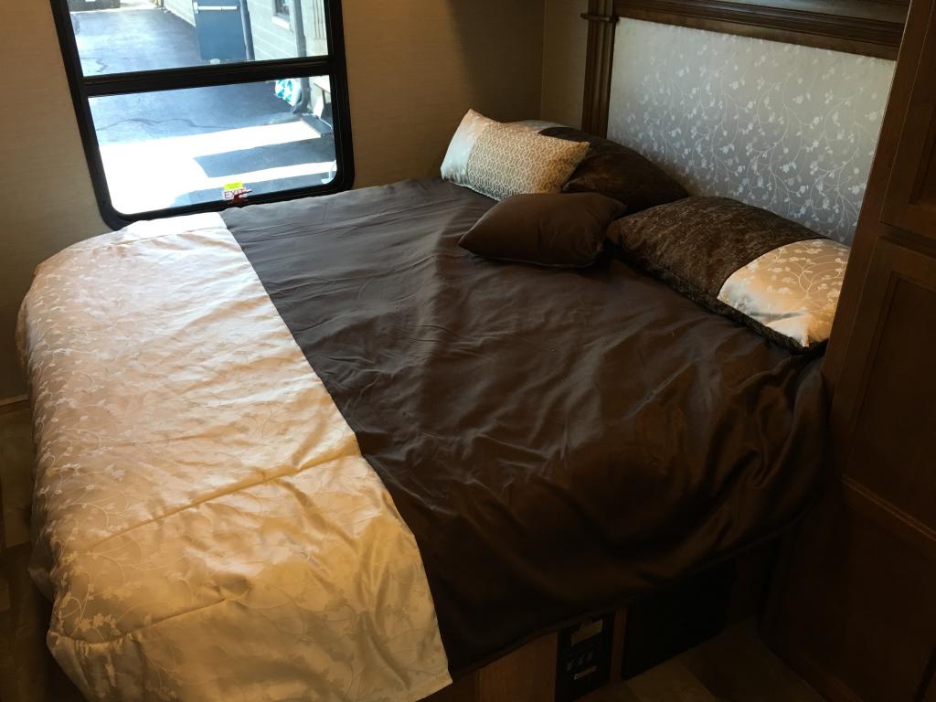 An RV rental bedroom features a comfy bed with dark sheets, a light comforter, and decorative pillows near a window.. Jayco Seneca 2019