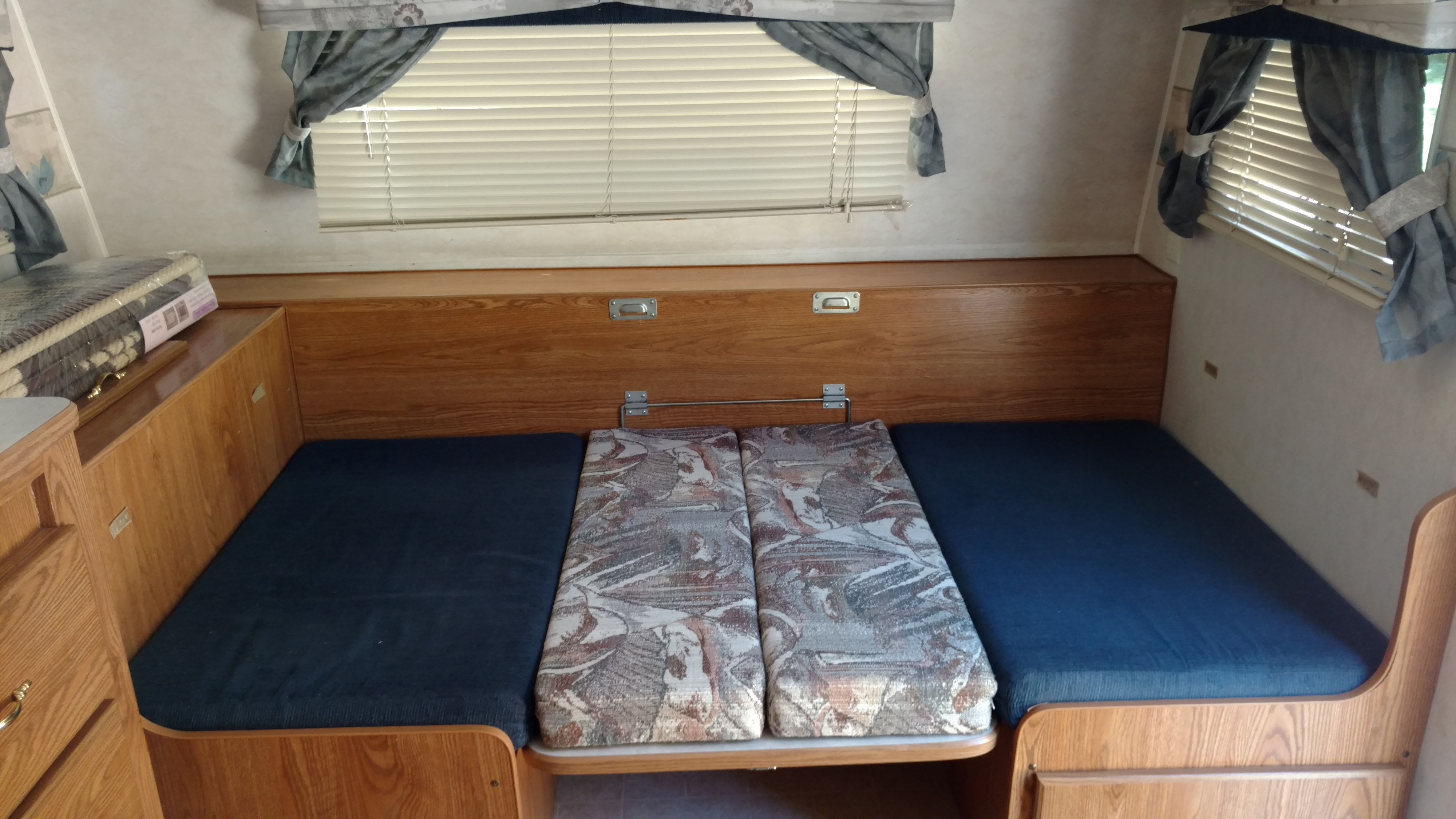 An RV rental interior with a convertible dinette and sleeping area. Features oak wood cabinetry, blue and patterned cushions, and a window.. Fleetwood Prowler 2002