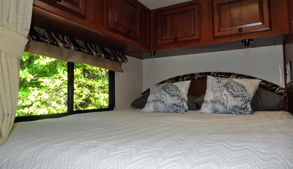 Cozy RV rental bedroom with wooden cabinets, white quilted bed, and pillows featuring owls, offering a view of lush greenery.. Forest River Coachmen Leprechaun 2014