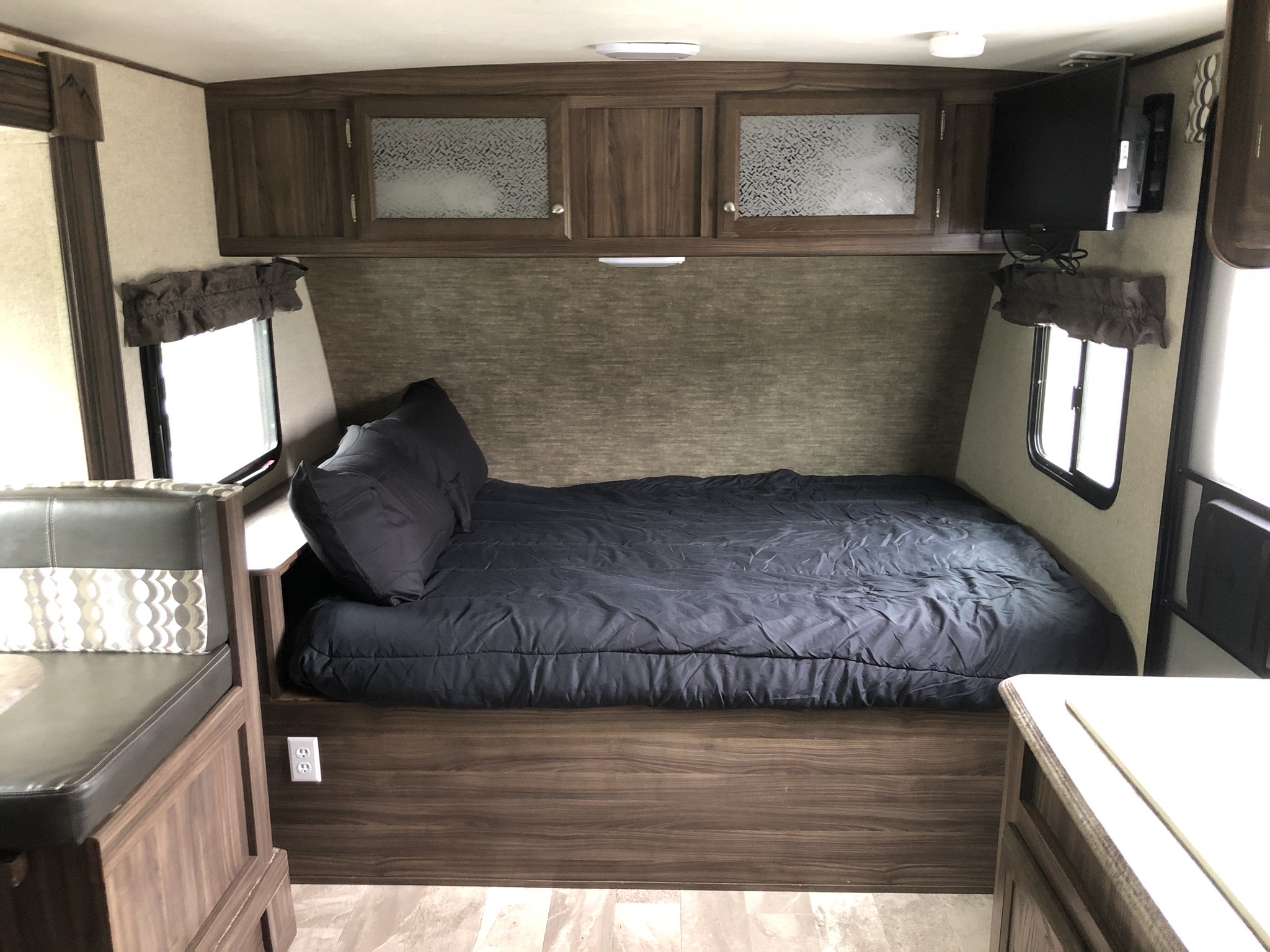 Modern RV rental interior showing a comfortable dark bed, overhead wood cabinets, mounted TV, and a dinette.. Coachmen Apex 2018