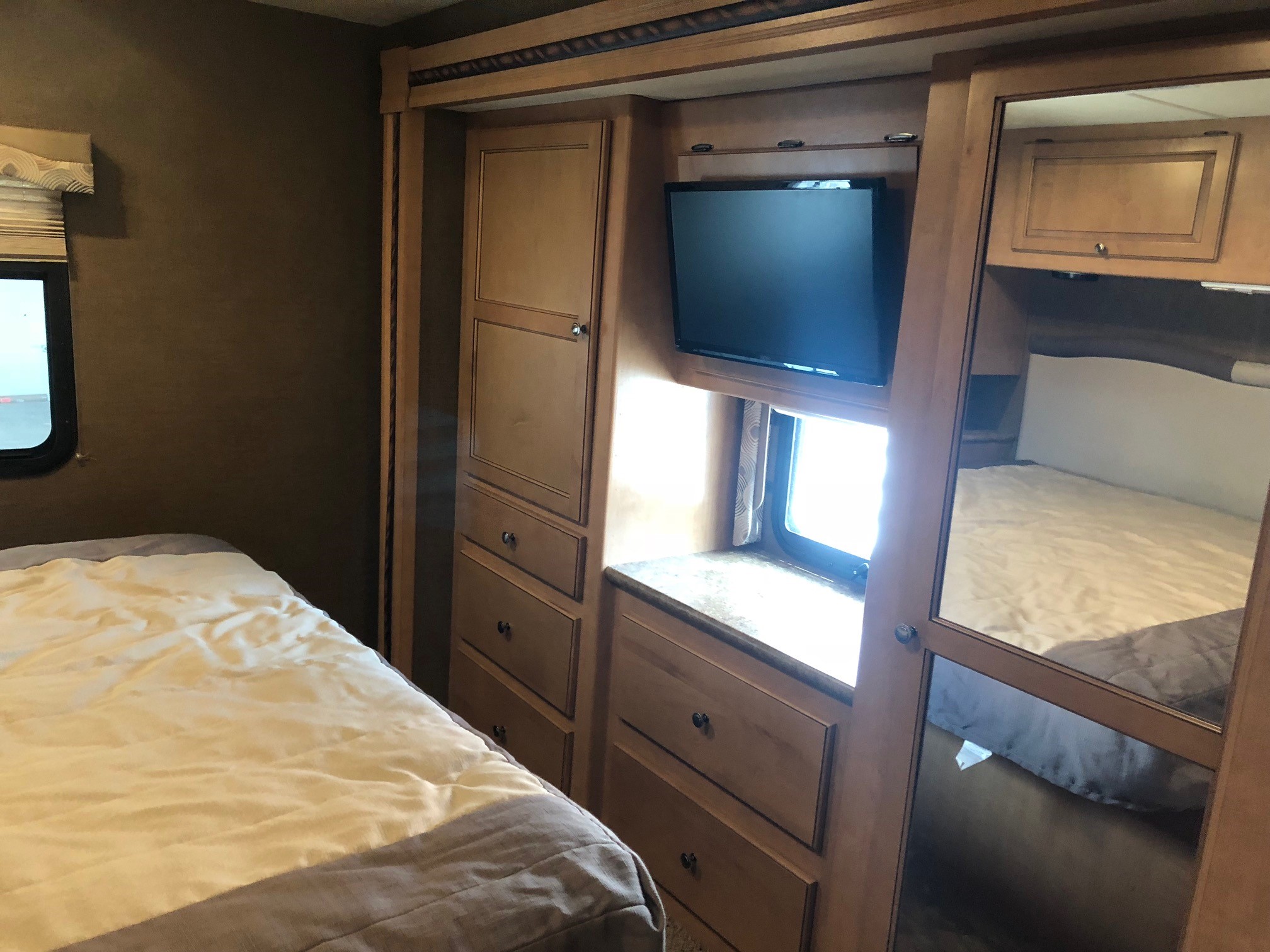 Inside an RV rental bedroom featuring a neatly made bed, wood cabinetry with drawers, a TV, and a mirrored closet.. Thor Motor Coach Windsport 2015