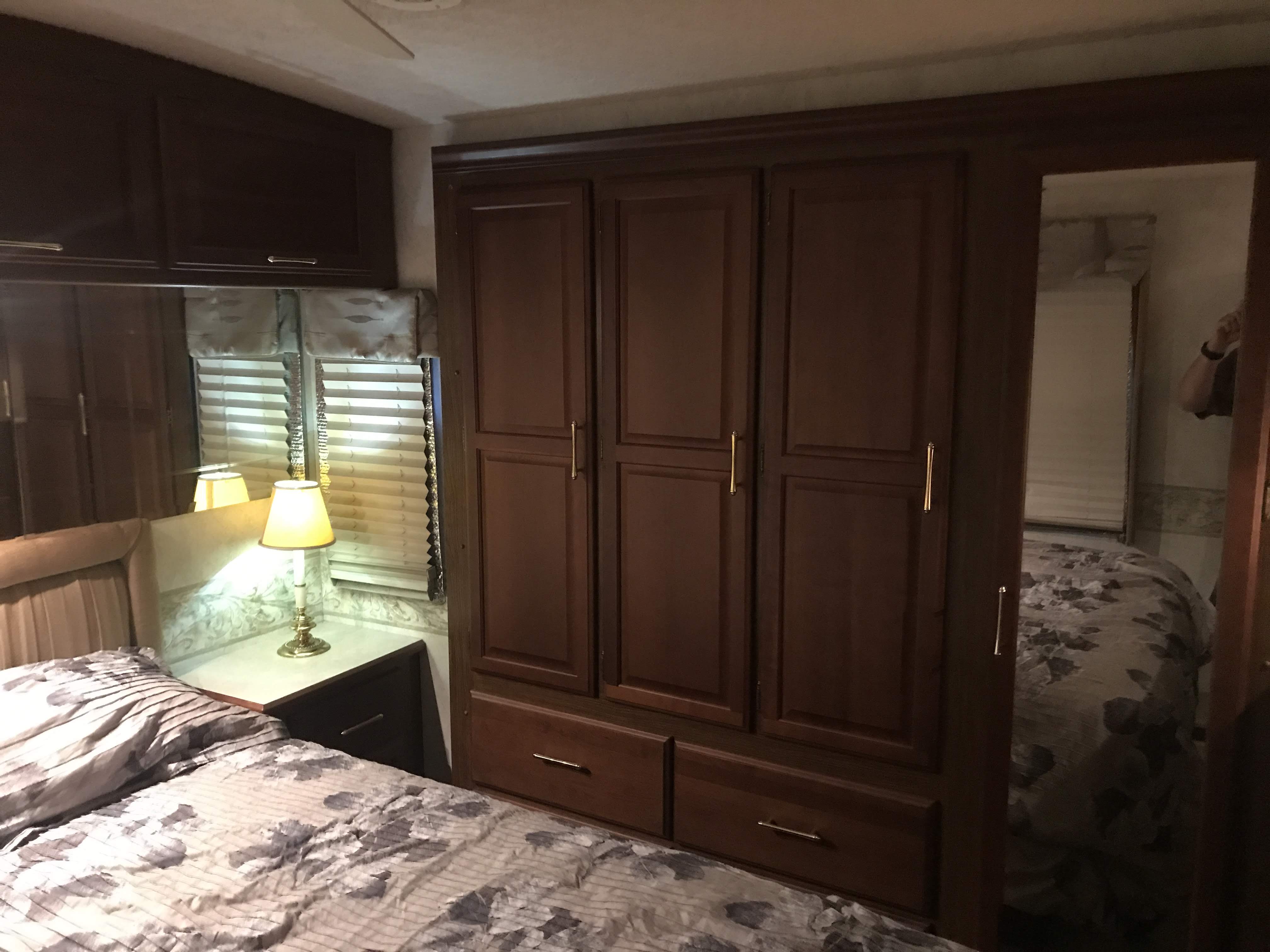 Cozy RV rental bedroom featuring a patterned bed, nightstand with lamp, and extensive built-in wooden cabinets. A mirror shows a reflection.. Winnebago Journey 2004