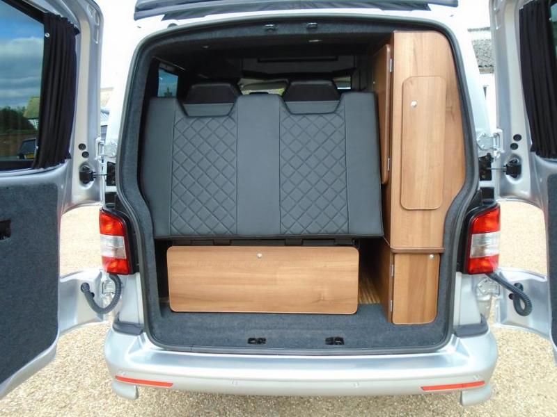 Rear view of a silver camper van's interior. Features a grey quilted foldable bed/seat and wooden storage units, ideal for an RV rental.. Volkswagen T5 premium camper 2012
