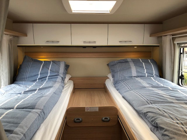 RV rental interior: cozy bedroom with two single beds, striped bedding, overhead storage, and warm ambient light.. Sunlight 2024
