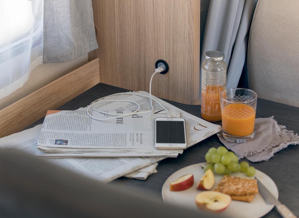 Cozy RV rental interior: A phone charges via a built-in USB, next to newspapers, juice, and a plate of fruit & crackers.. Sunlight T69LC 2024