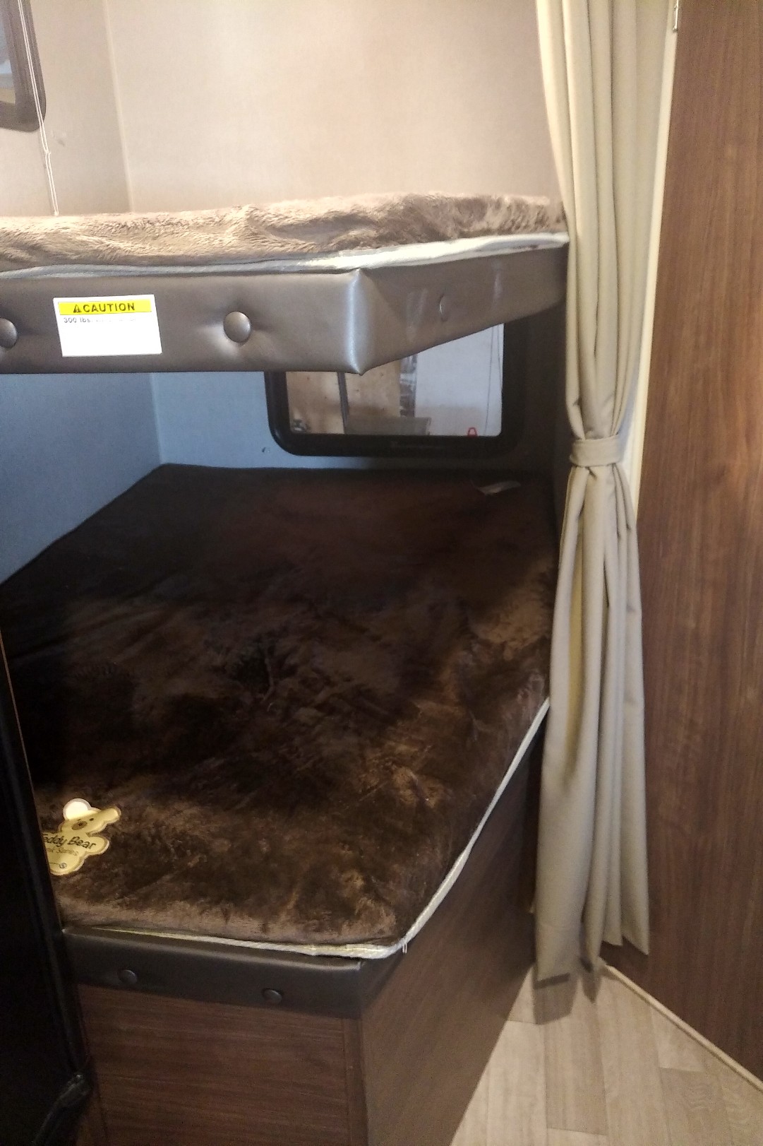 Inside an RV rental: two dark brown, plush bunk beds stacked. A "300 lbs" caution sticker is on the upper bunk, with a tied-back curtain to the right.. Coleman 244BH 2017