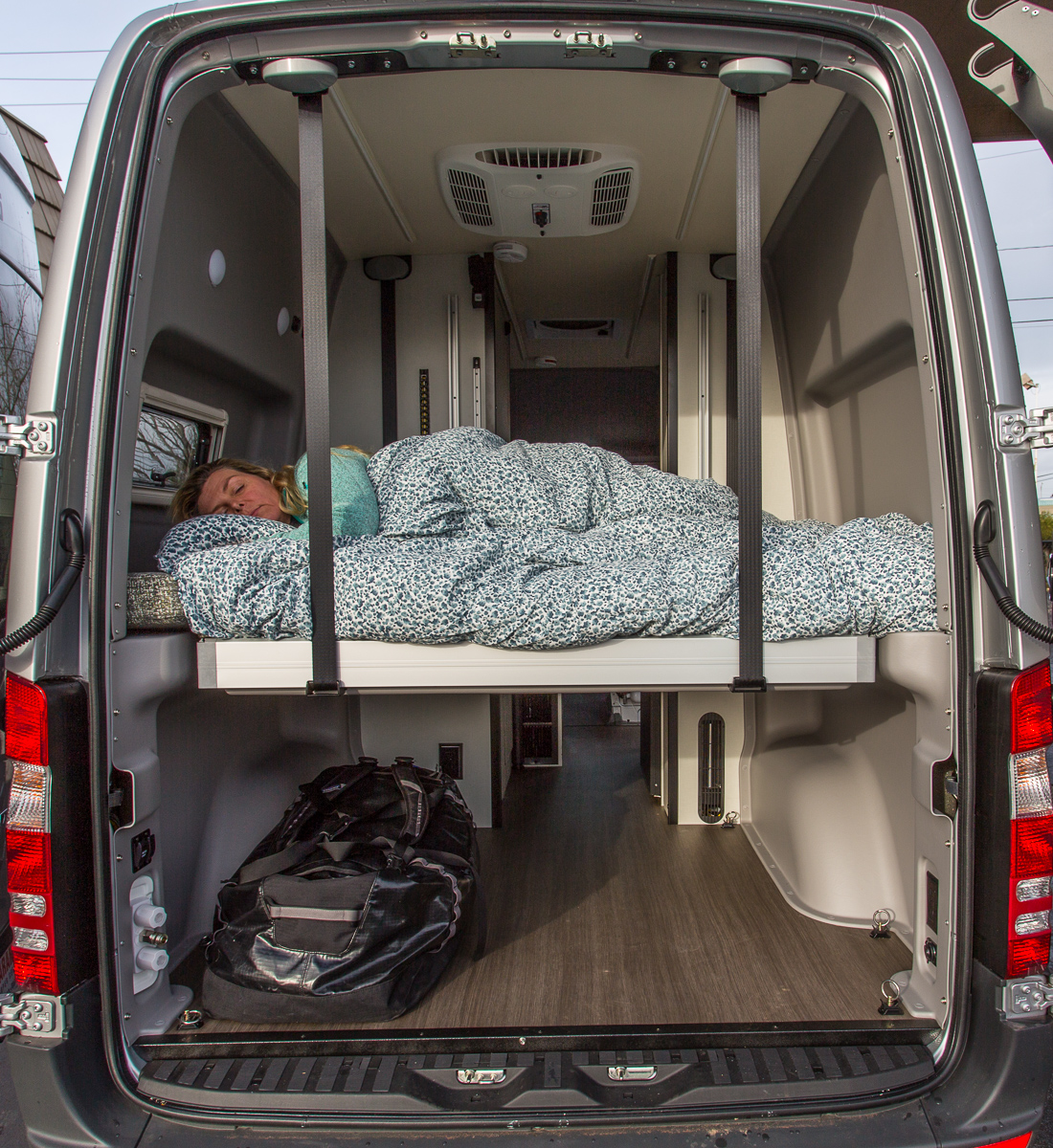 RV rental camper van interior: A person sleeps on a raised bed. Below, ample storage space includes a duffel bag.. Winnebago Other 2018
