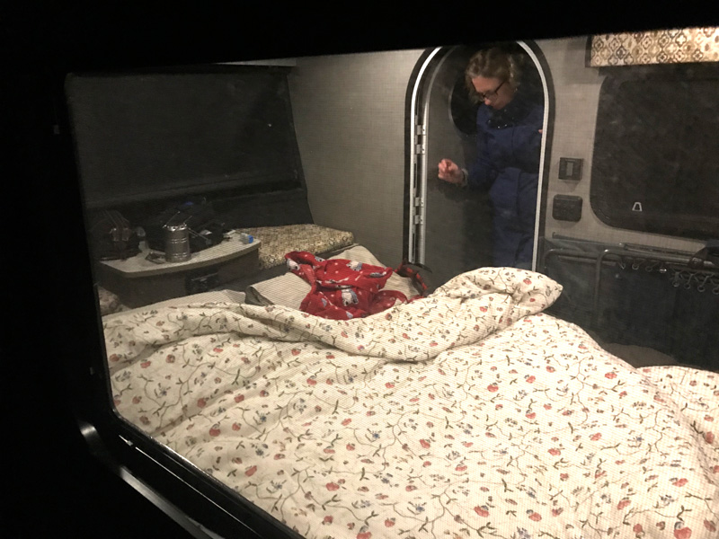 Relaxing RV rental interior with floral bed covers, viewed through a screen. A person in blue stands by the doorway.. InTech RV Luna 2018