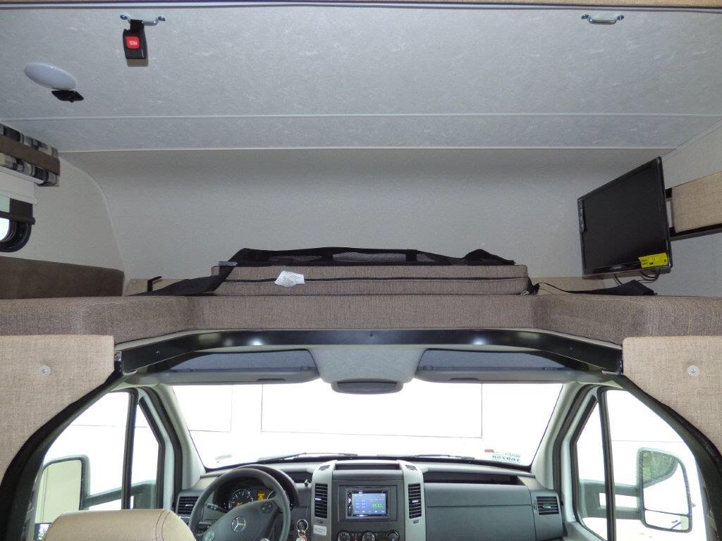 An RV rental interior, from the living space, looking towards the driver's cab below an overhead bunk bed with a wall-mounted TV.. Other Other 2018