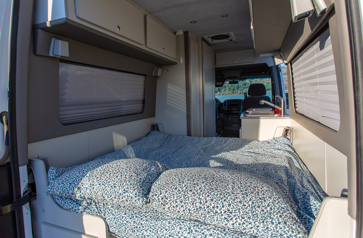 RV rental interior with a bed, blue patterned bedding, overhead cabinets, and a compact kitchen with a sink.. Freightliner Sprinter 2011
