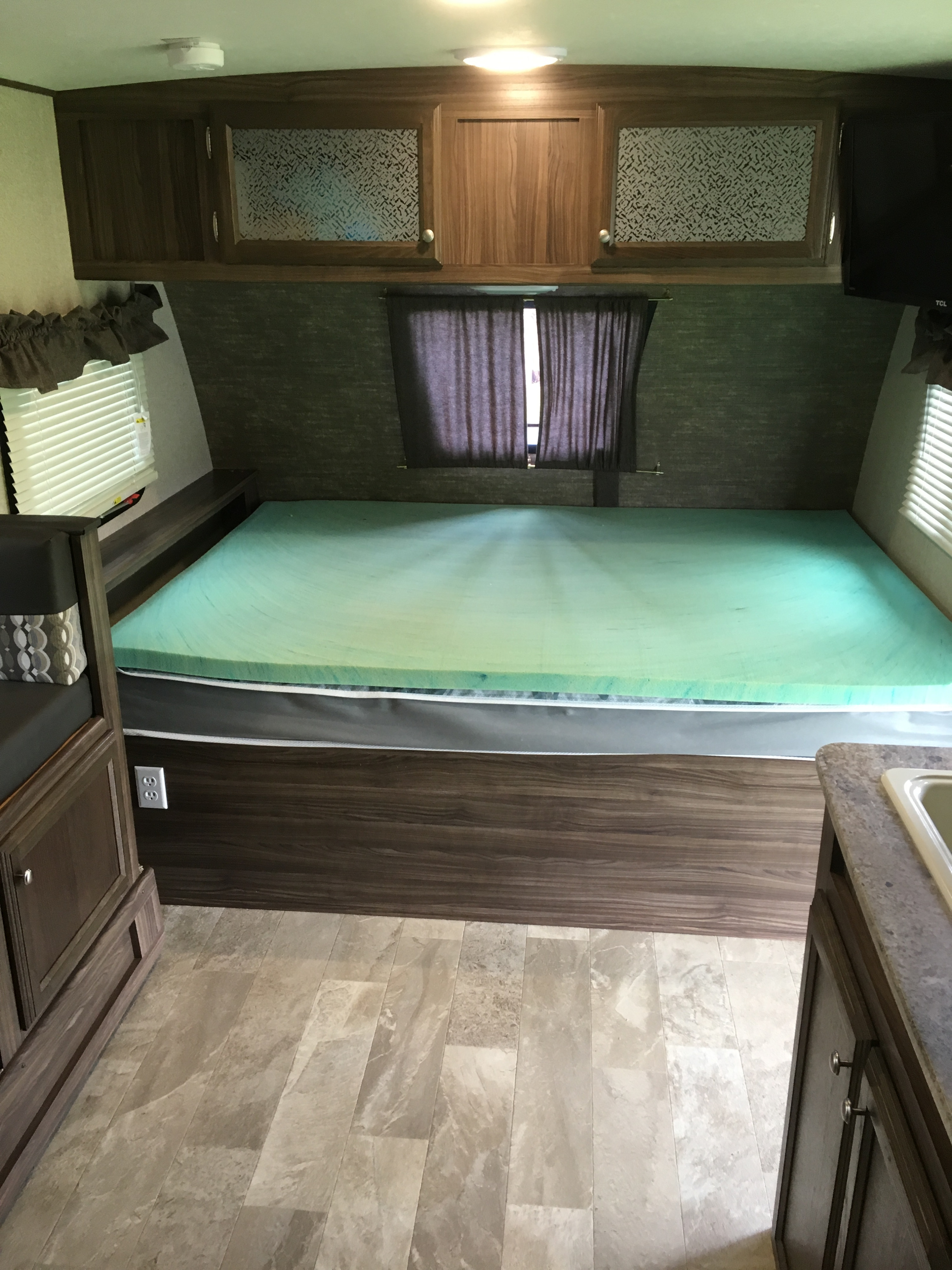 RV rental interior featuring a cozy bed with a green topper, overhead cabinets, and partial kitchen area.. Coachmen Apex 2018