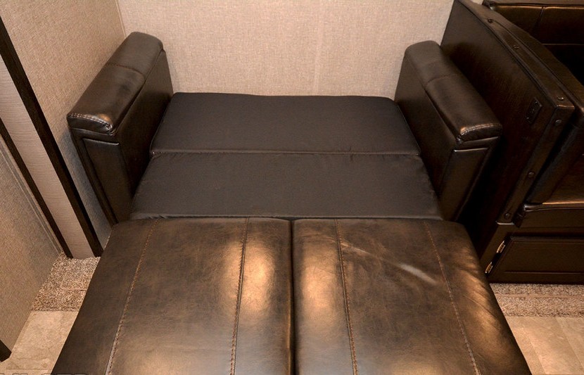RV rental comfort: A brown convertible sofa fully extended as a bed, showing the flat sleeping surface in a cozy space.. Grand Design Imagine 2019