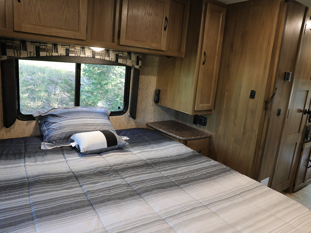 Comfortable bedroom interior in an RV rental, featuring striped bedding, cabinets, and a window with a view. Perfect for camping trips.. Ford Coachman 2018