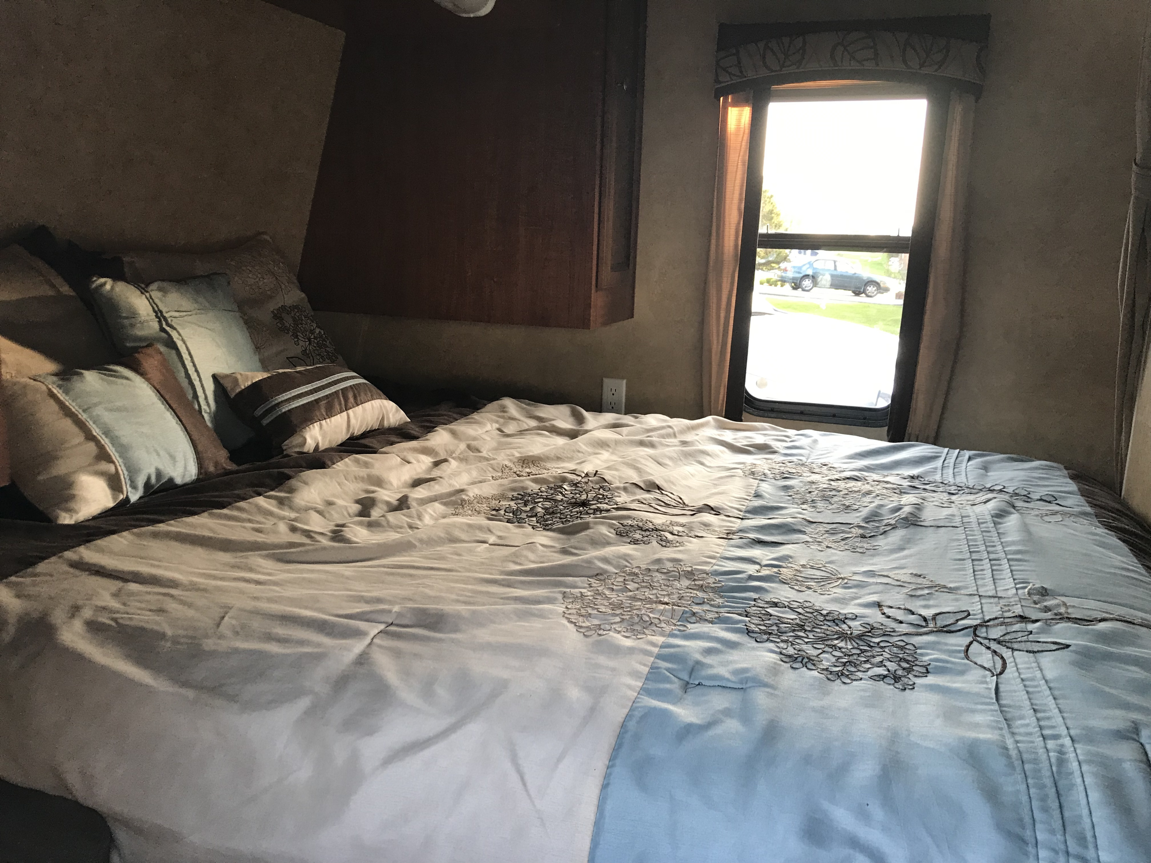Cozy RV rental bedroom with patterned comforter and pillows, offering a bright window view of the outdoors and a parked car.. Jayco Jay Flight 2012