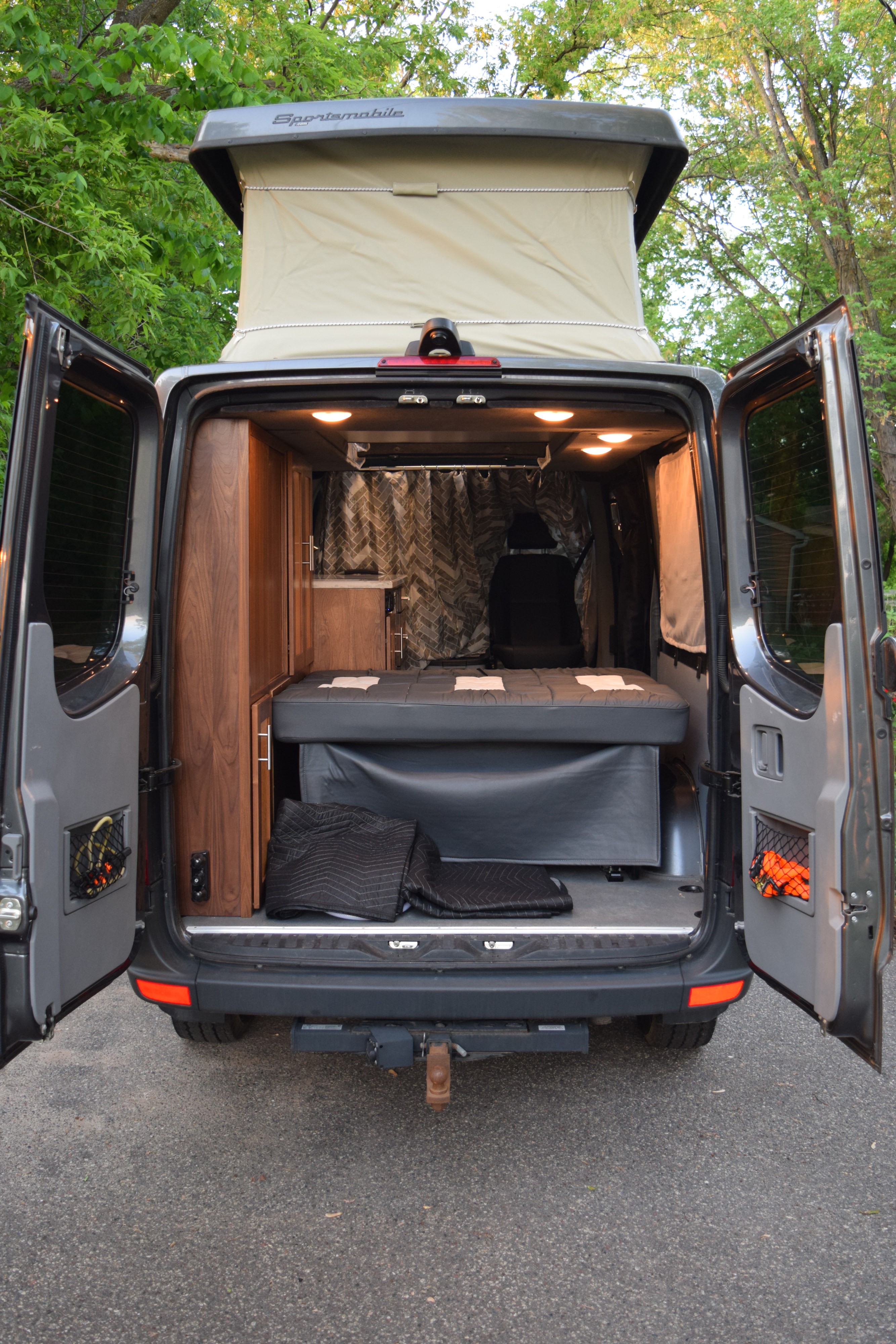 Experience this custom van RV rental! Rear view reveals open doors, pop-top roof, bed, and warm wood interior.. Mercedes-Benz Sprinter 2016