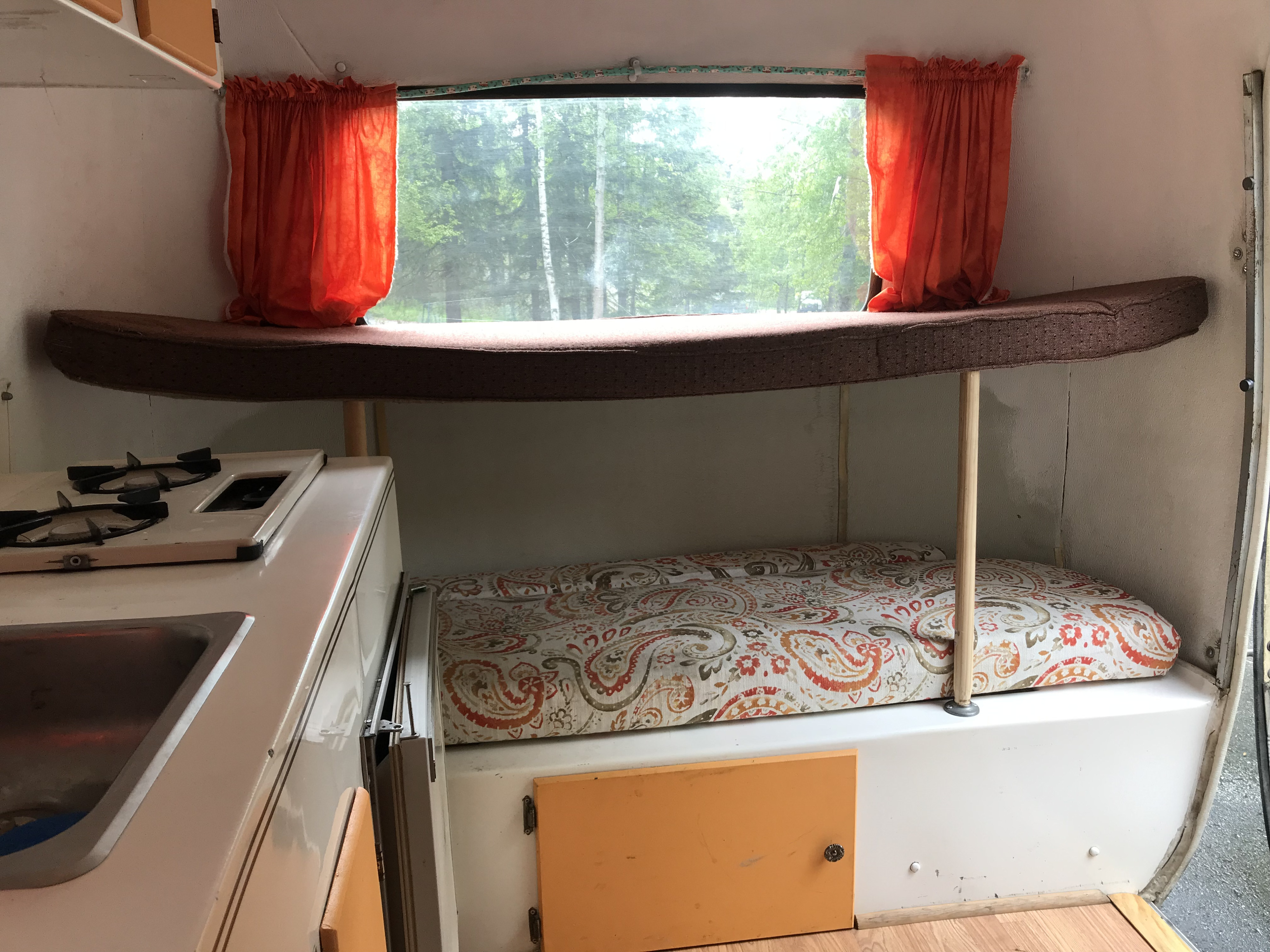 Inside a cozy RV rental: bunk beds, kitchenette with sink/stove, and a scenic window view of green trees.. Scamp 13' 1987