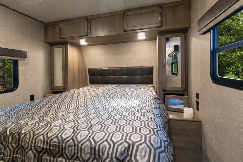 This RV rental bedroom offers a comfortable bed with a padded headboard, overhead storage, and scenic window views.. Grand Design Transcend 2020