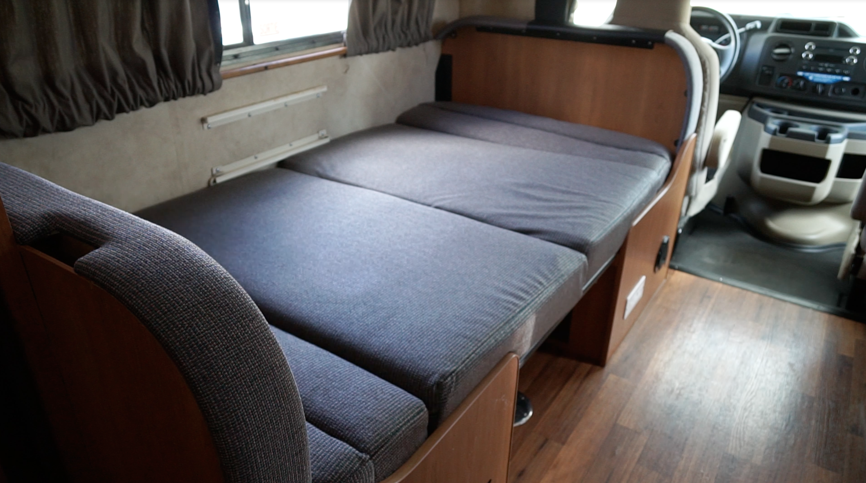 Grey cushions convert into a bed inside this RV rental, with wood-look flooring & the driver's cabin visible.. Majestic Leisure Craft 23A 2014
