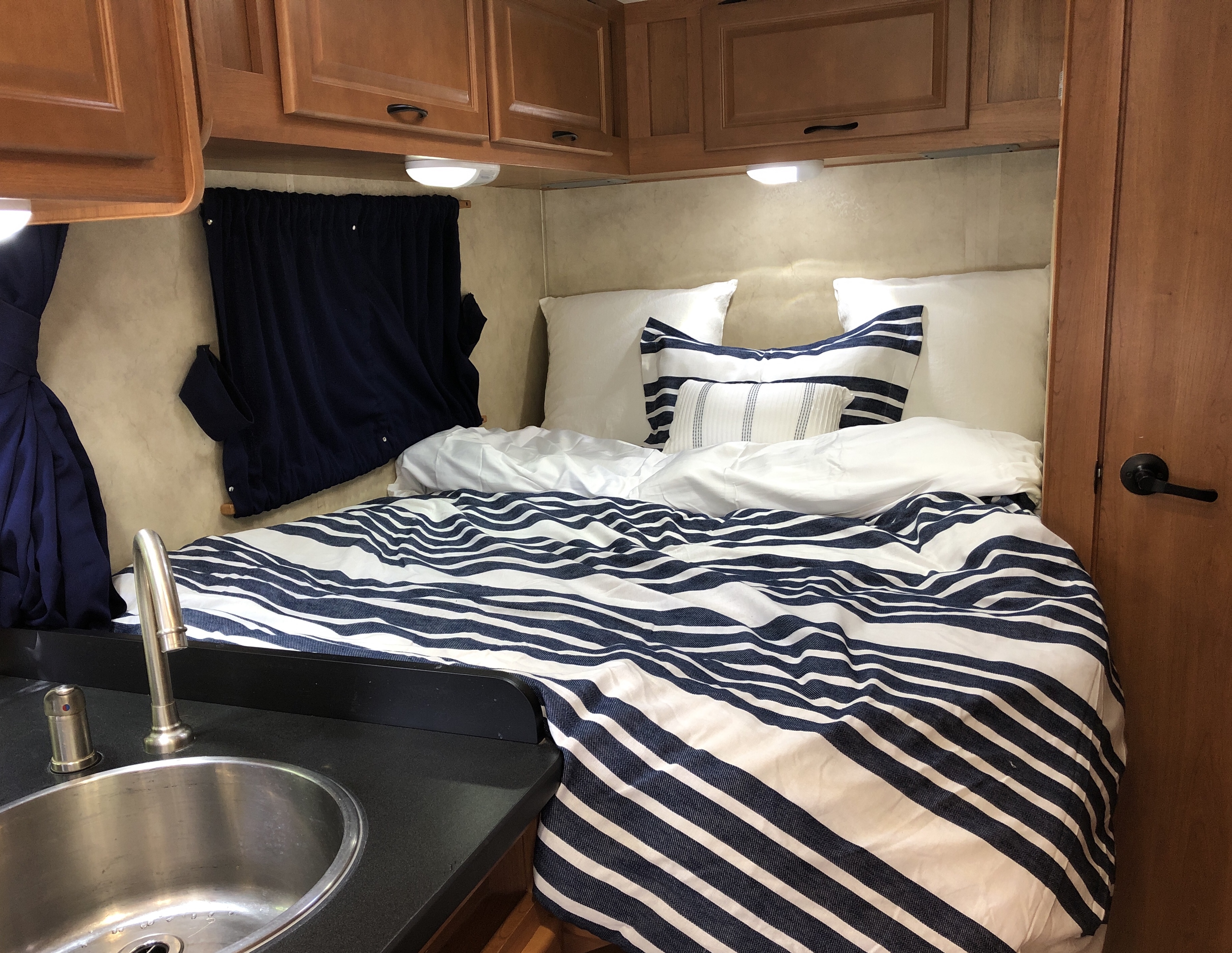 Cozy RV rental bedroom with striped bedding, overhead cabinets, and a compact kitchen sink on the left.. Thor Majestic 2014