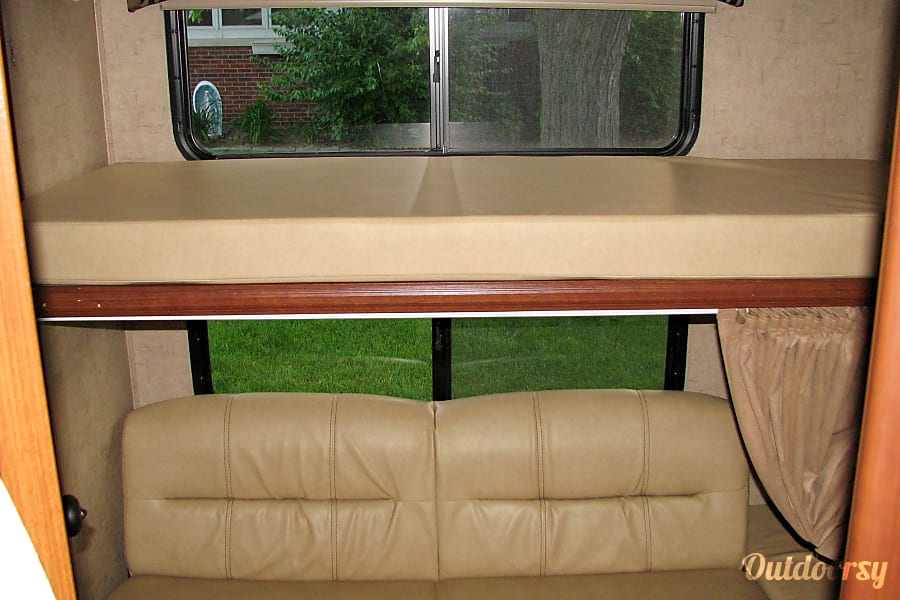 RV rental interior view shows a beige bunk bed over a matching sofa. Windows overlook green outdoors.. Forest River Sunseeker 2015