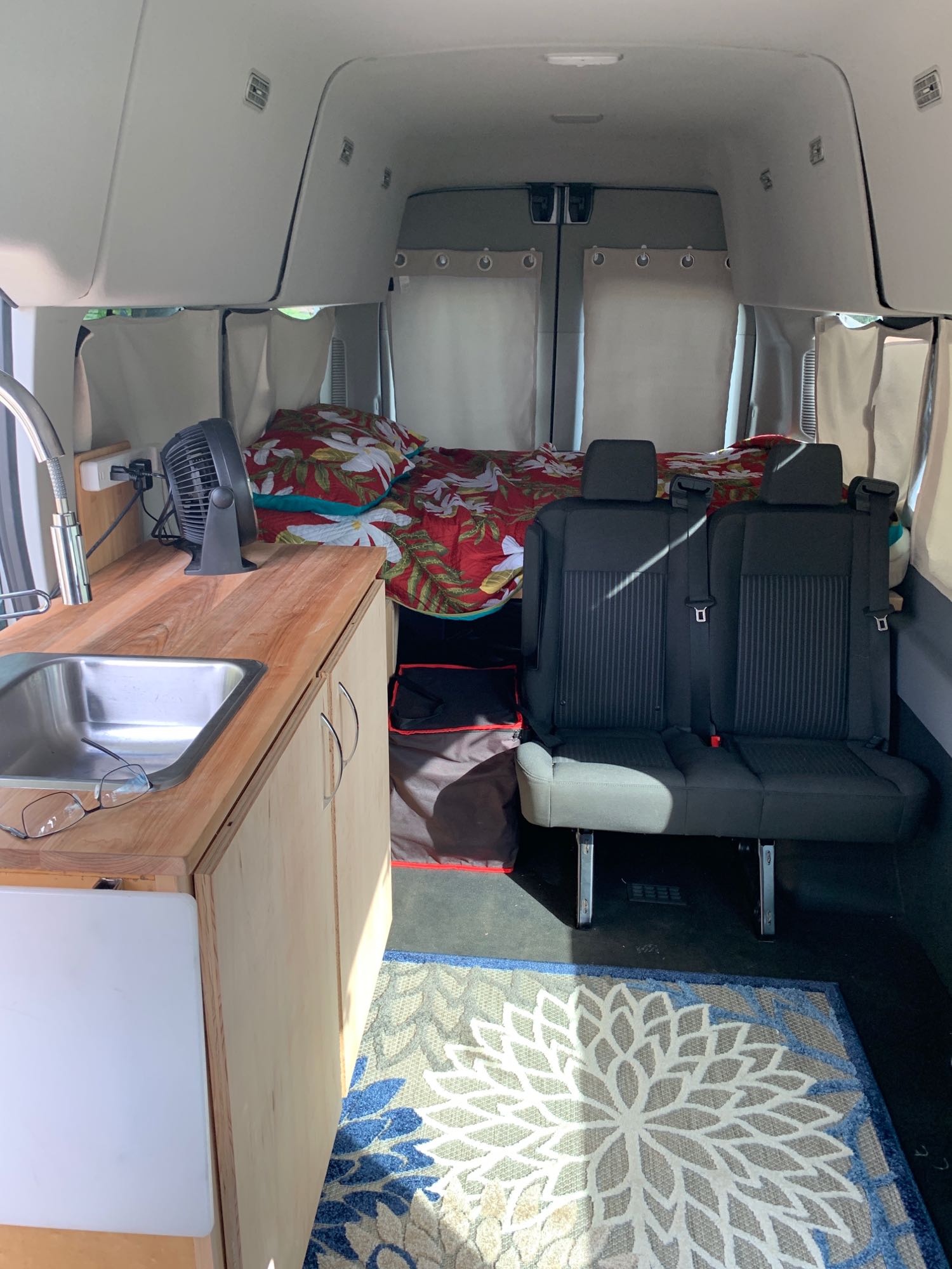 Interior of an RV rental camper van, featuring a kitchen counter with sink, cozy bed with floral bedding, and two passenger seats.. Ford Transit 2018