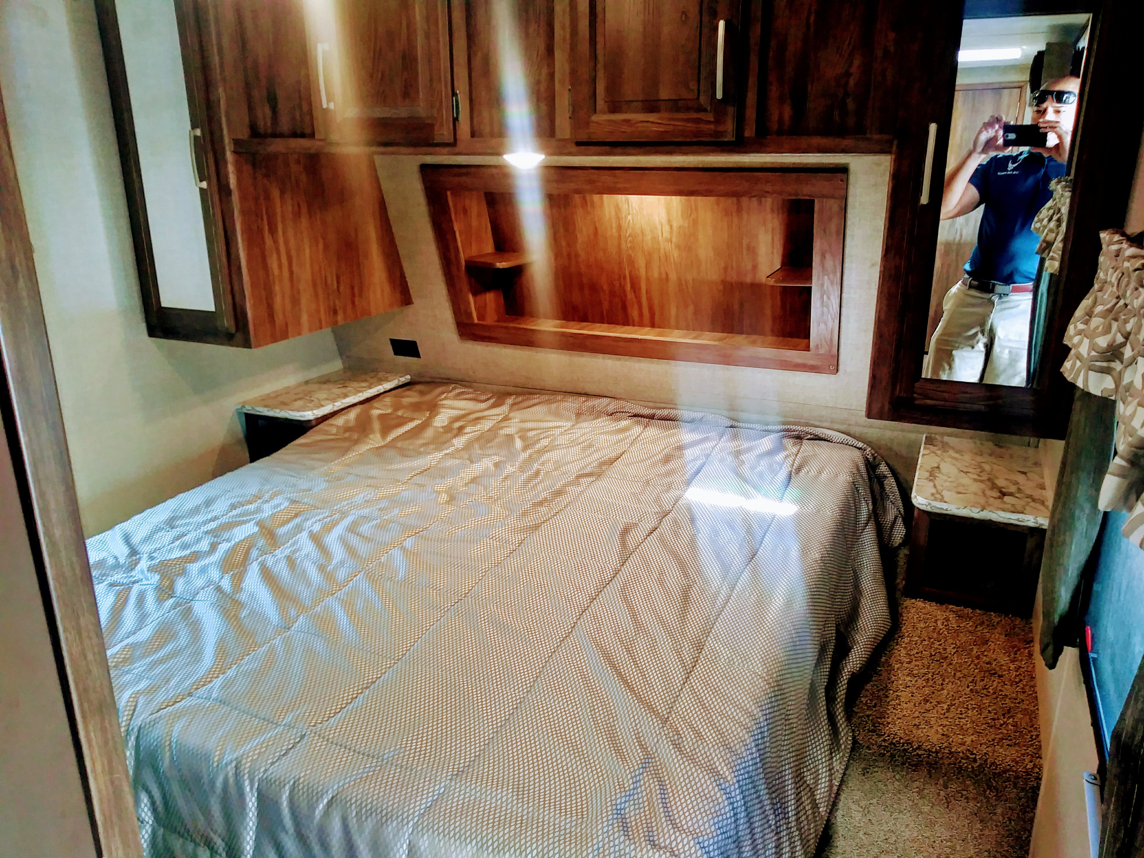 RV rental bedroom featuring a patterned bedspread, wooden cabinets, and a mirror reflecting the photographer.. Keystone Cougar Lite 2017