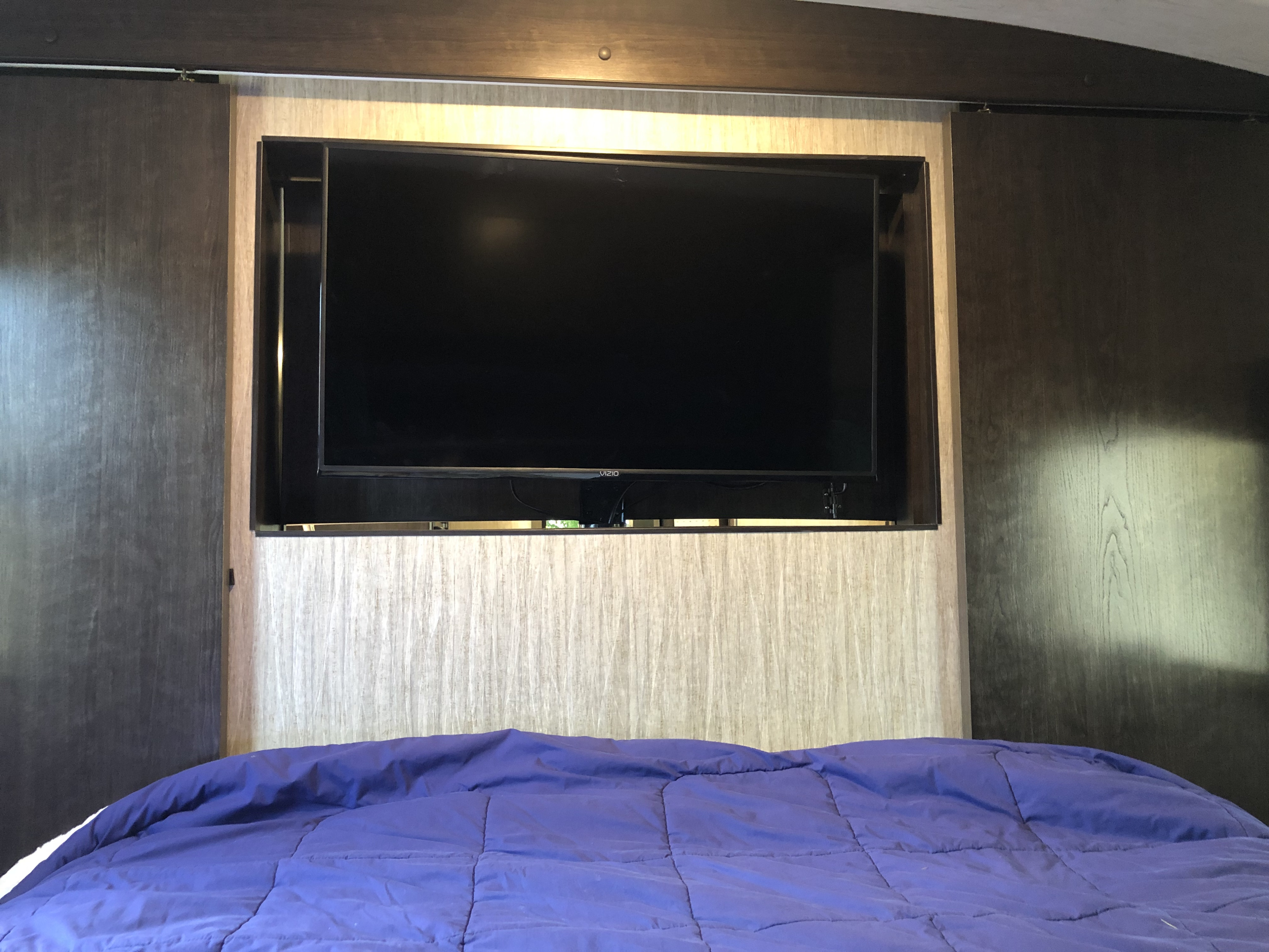 Inside this RV rental bedroom: a mounted TV above a purple bed, flanked by dark sliding doors. Ideal for cozy travels.. Heartland Sundance 2019
