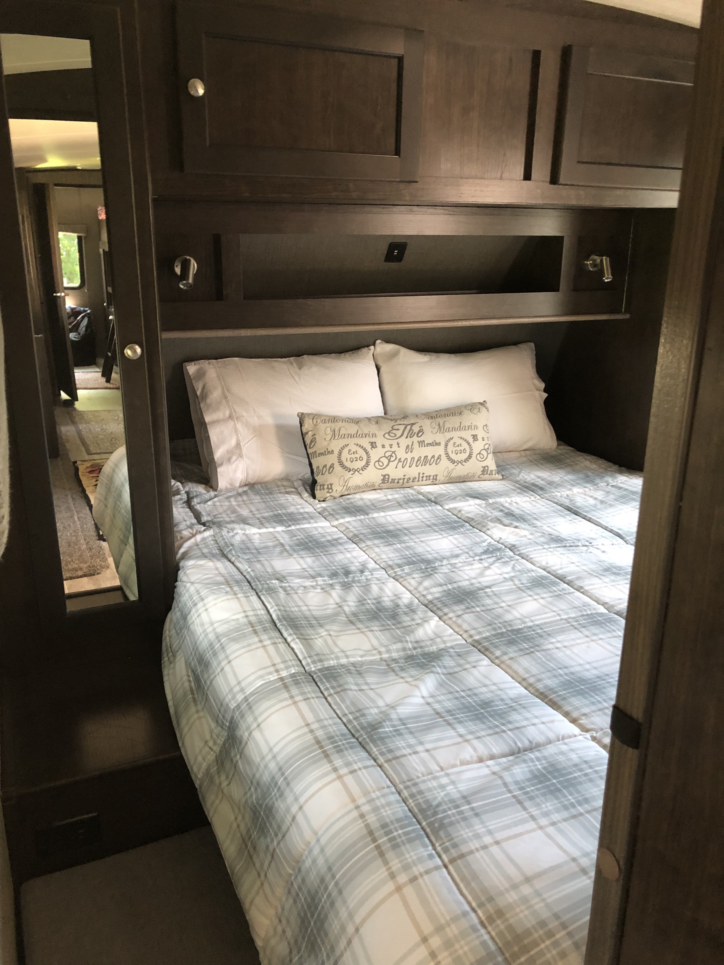 Cozy RV rental bedroom with dark wood cabinets, a plaid bed, and a mirrored closet reflecting the hallway.. Heartland Sundance 2019