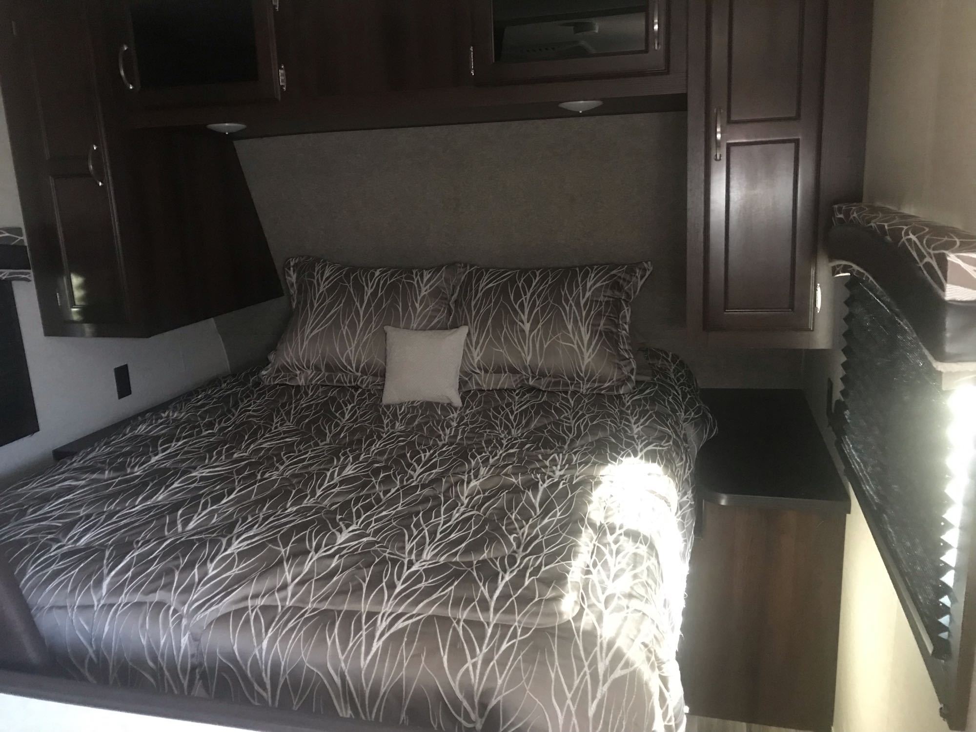 Cozy RV rental bedroom featuring a comfortable bed with patterned bedding, pillows, overhead cabinets, and a nightstand.. Jayco Jay Flight 2018