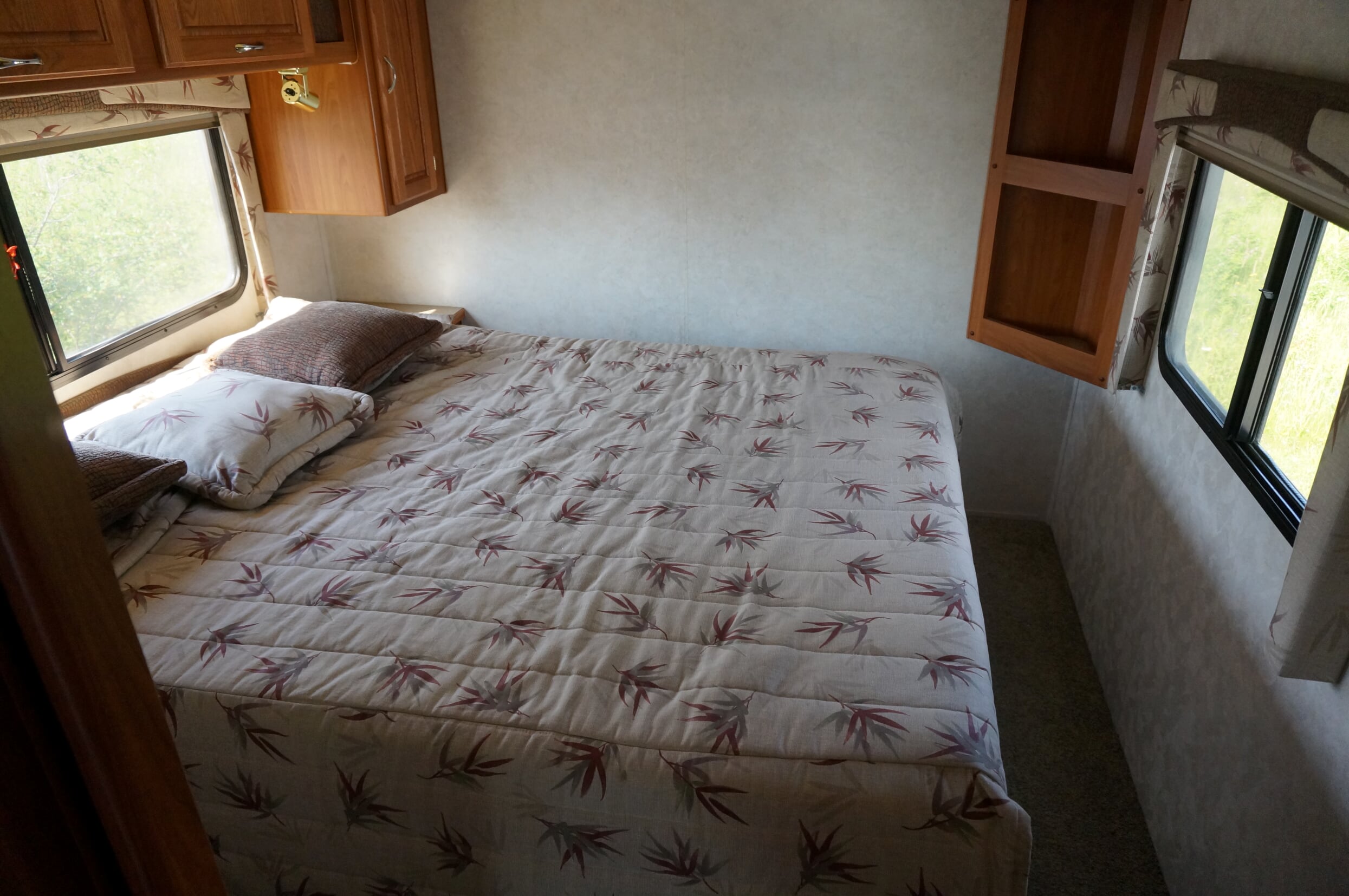 Compact RV rental bedroom with a comfortable bed, overhead cabinets, and bright windows displaying outdoor greenery.. Bigfoot 30MH29G 2004