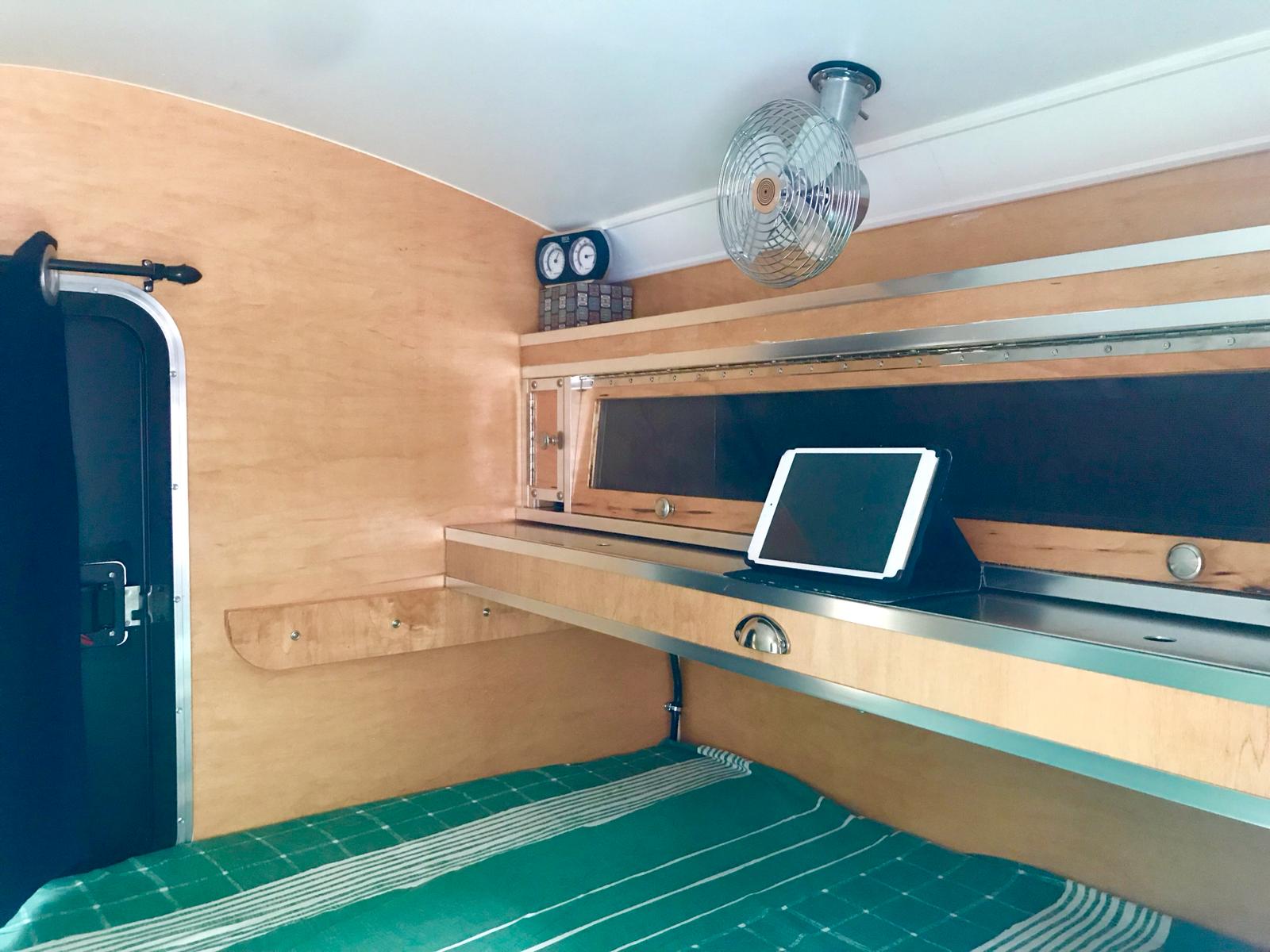 Step inside this cozy RV rental with light wood interior, green bedding, and fan. Tablet on shelf, compact camper living.. The Teardrop Trailer. N/A 2019