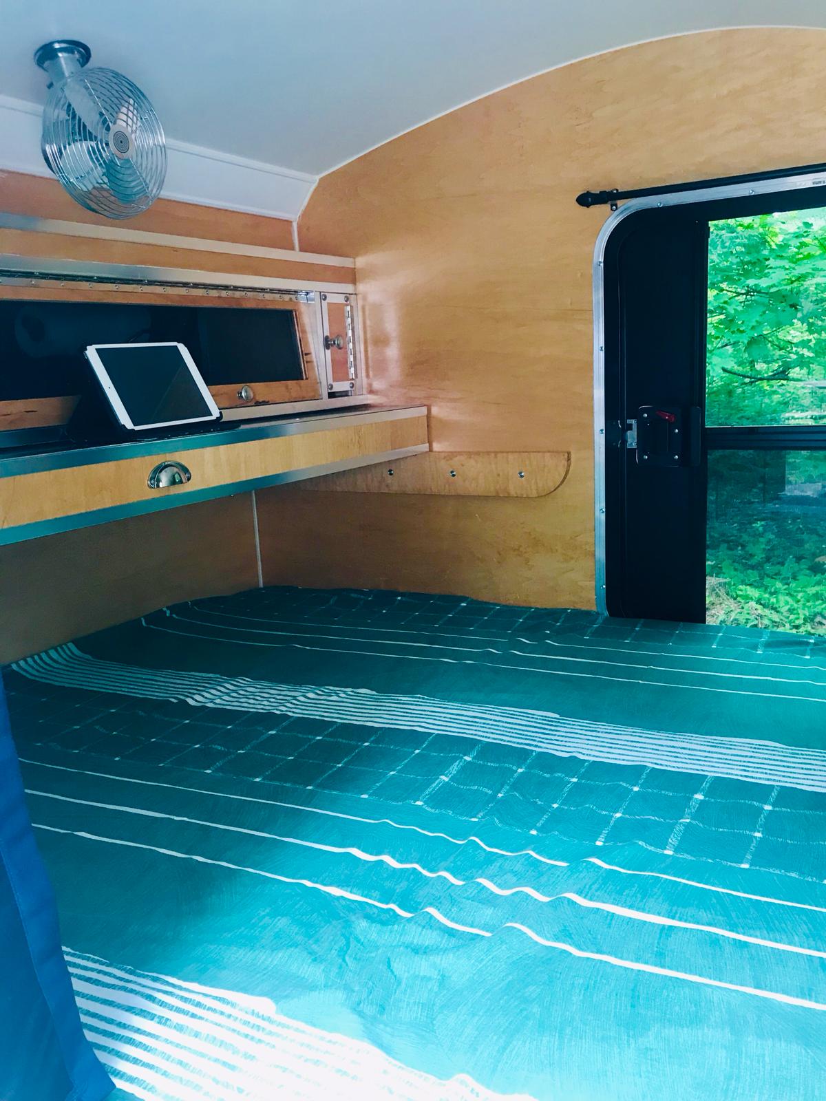 Cozy RV rental interior showcasing a bed, overhead fan, and an iPad on a shelf. Ready for peaceful travels amidst nature.. The Teardrop Trailer. N/A 2019