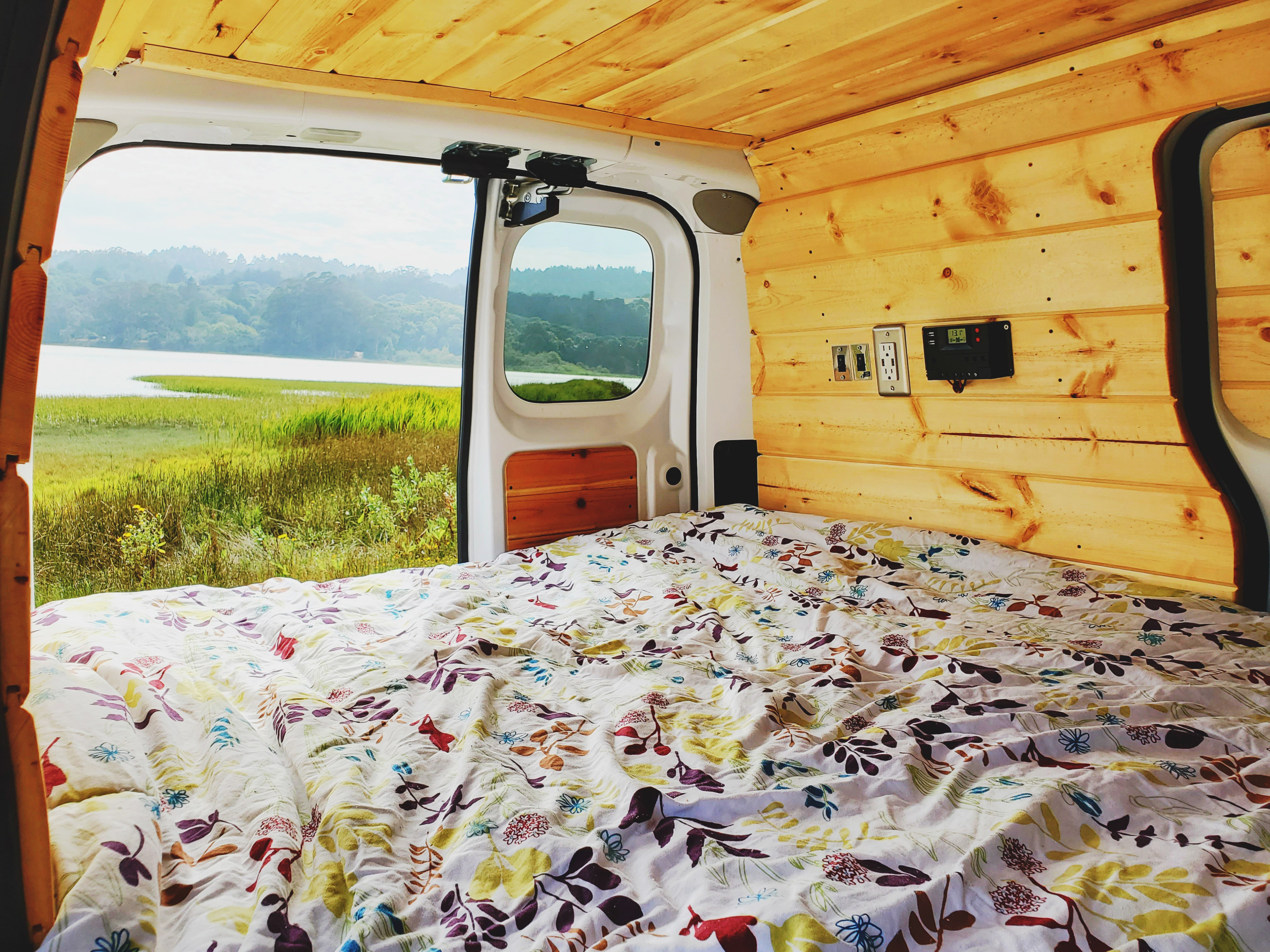 RV rental view: Cozy van interior with wooden walls, floral bed, and stunning lake landscape out the back doors.. Nissan NV200 2019